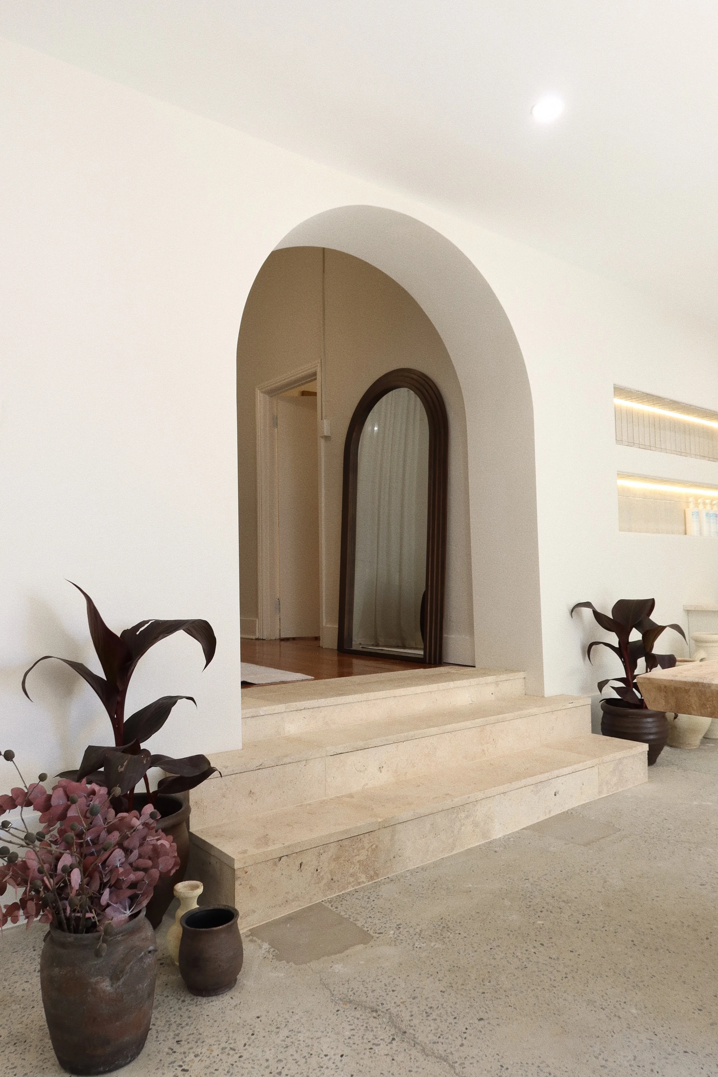 Minimalist indoor entrance with beige steps, potted plants, dark wooden framed mirror, and arched doorway.