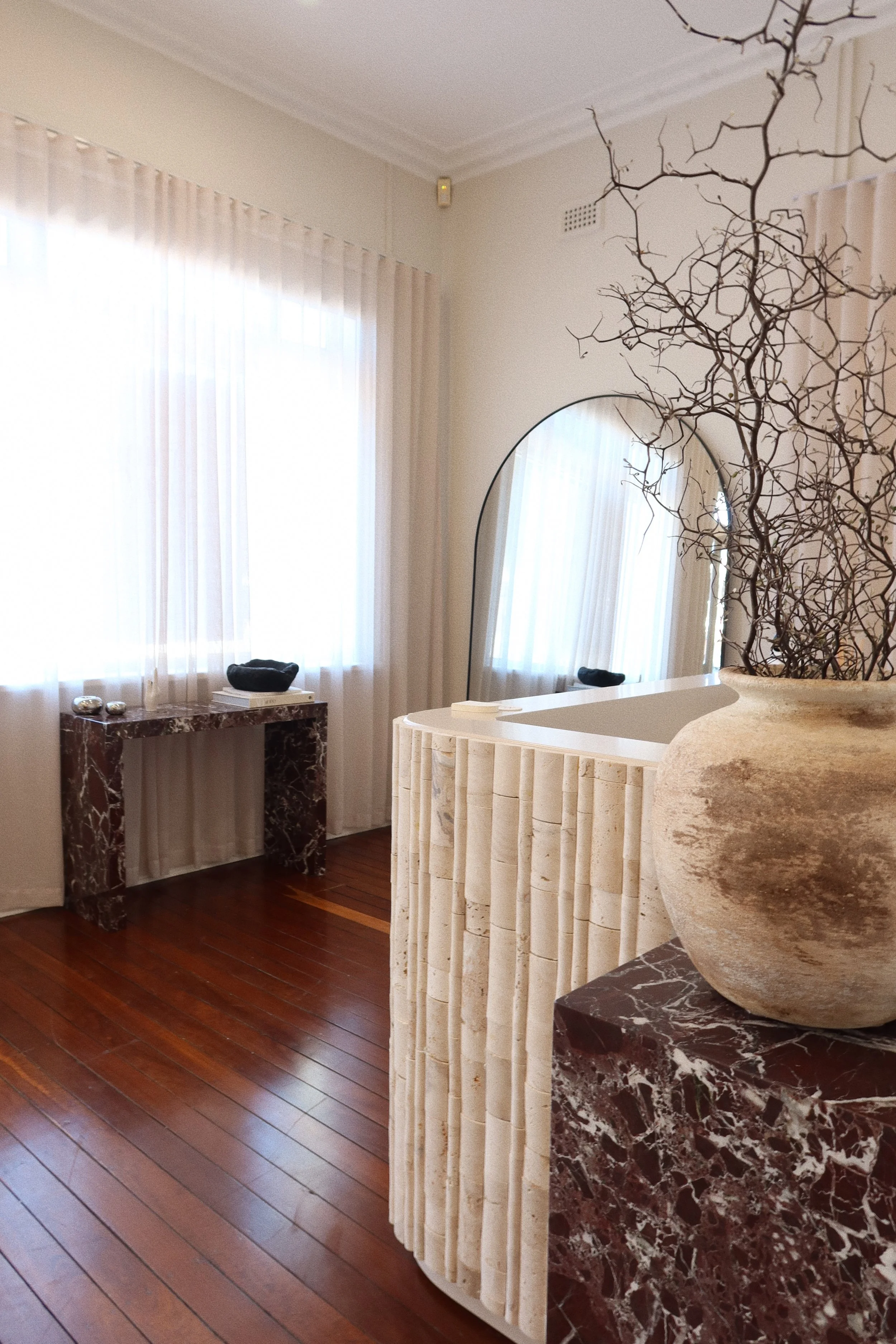 Interior of a room with sheer white curtains, a marble console table, a large oval mirror, and a decorative urn with branches, featuring warm wooden floors.