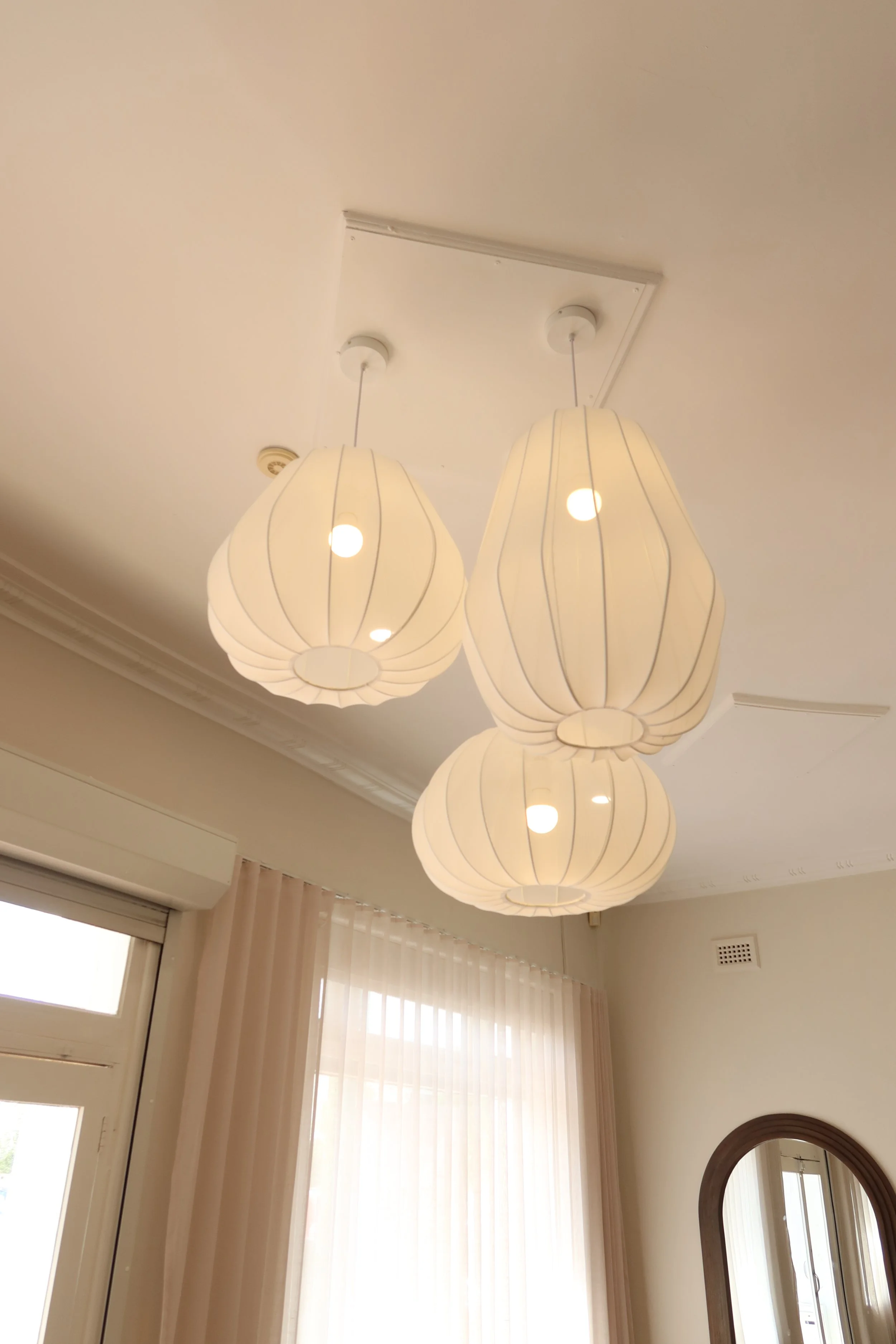 Overhead light fixture with three white paper lantern-style lampshades hanging from the ceiling in a well-lit room.