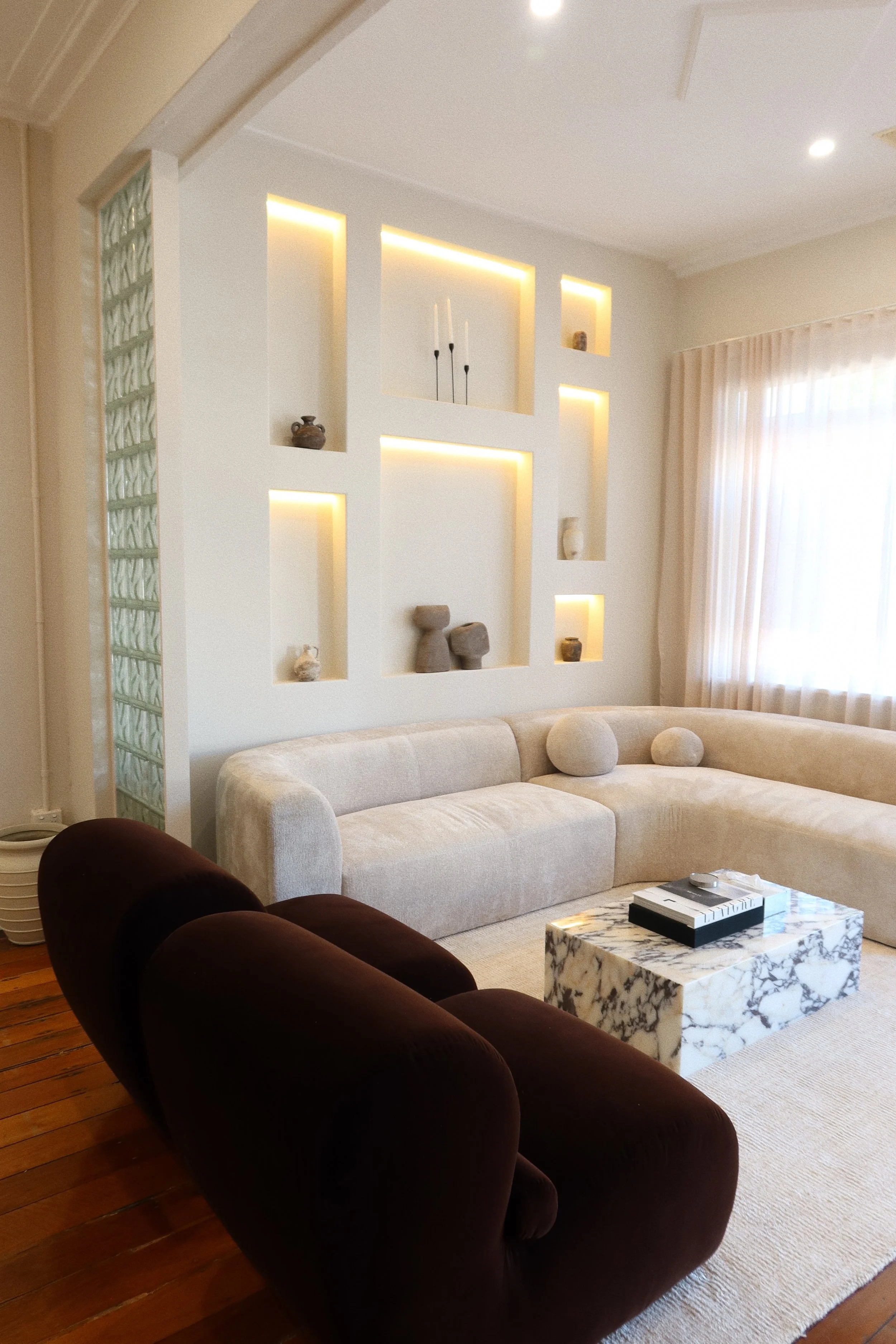 Living room with a white sectional sofa, two round pillows, dark velvety armchairs, a marble coffee table, and built-in wall shelves with decorative objects and lighting.