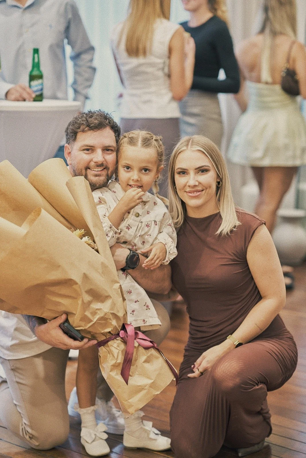A happy family, with a man holding a bouquet of flowers and a young girl, and a woman kneeling next to them, all smiling at the camera at a social gathering.