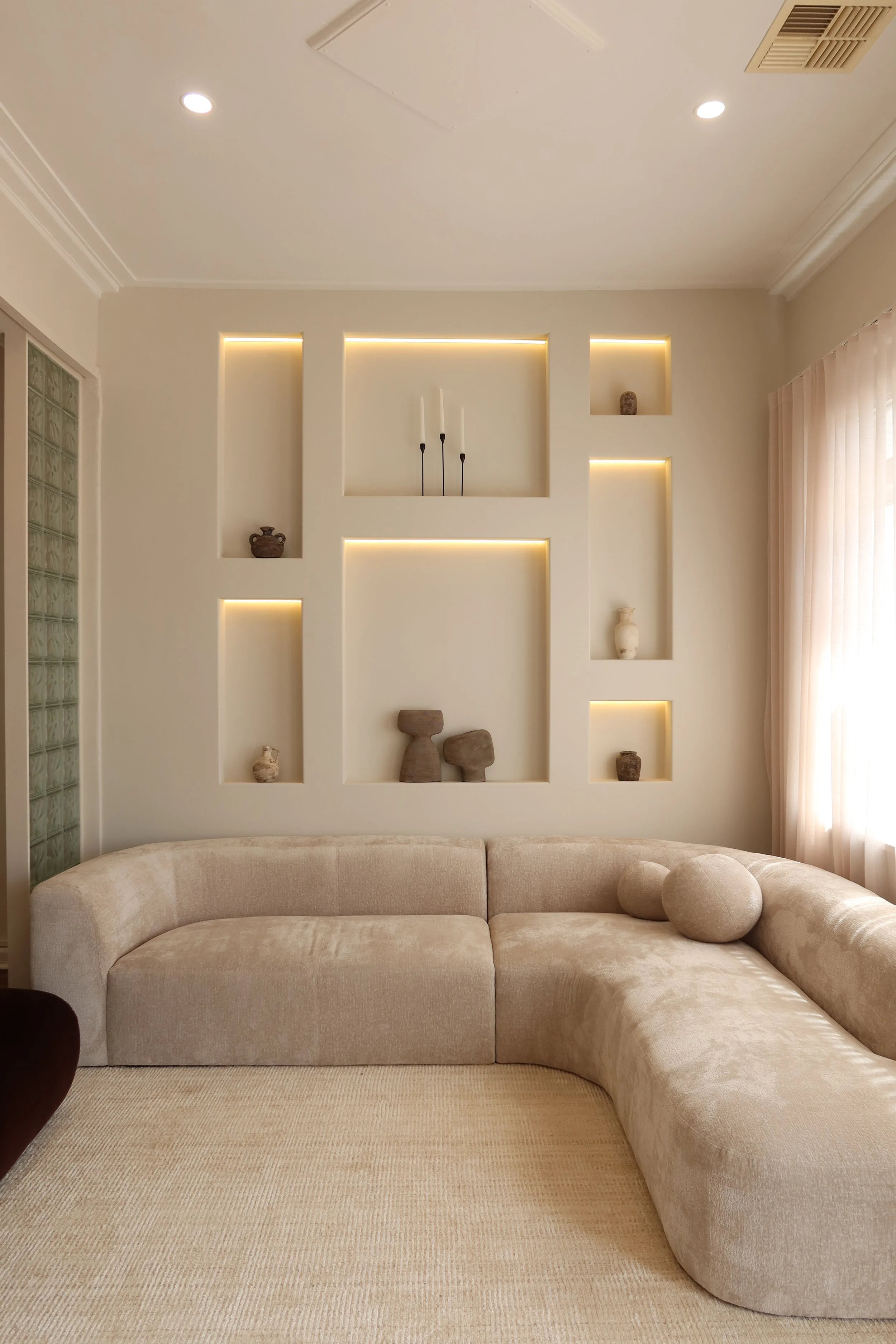 A beige sectional sofa with two round cushions, in front of a wall with built-in shelves illuminated by yellow lighting, holding various small decorative vases and sculptures. A window with sheer curtains is on the right.