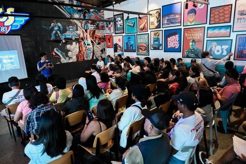 A large group of people attending a presentation in a crowded room with colorful posters on the wall featuring pop culture, artwork, and advertisements.