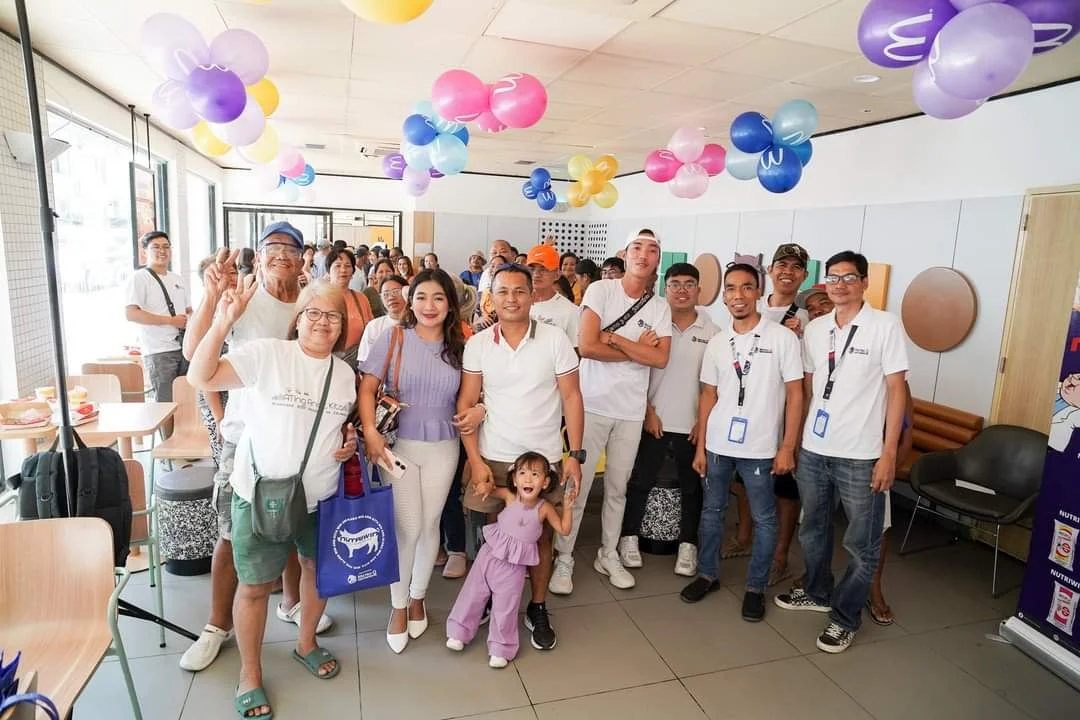 Group of people celebrating indoors with colorful balloons hanging from the ceiling, posing for a photo, some smiling and waving, in a bright and lively environment.