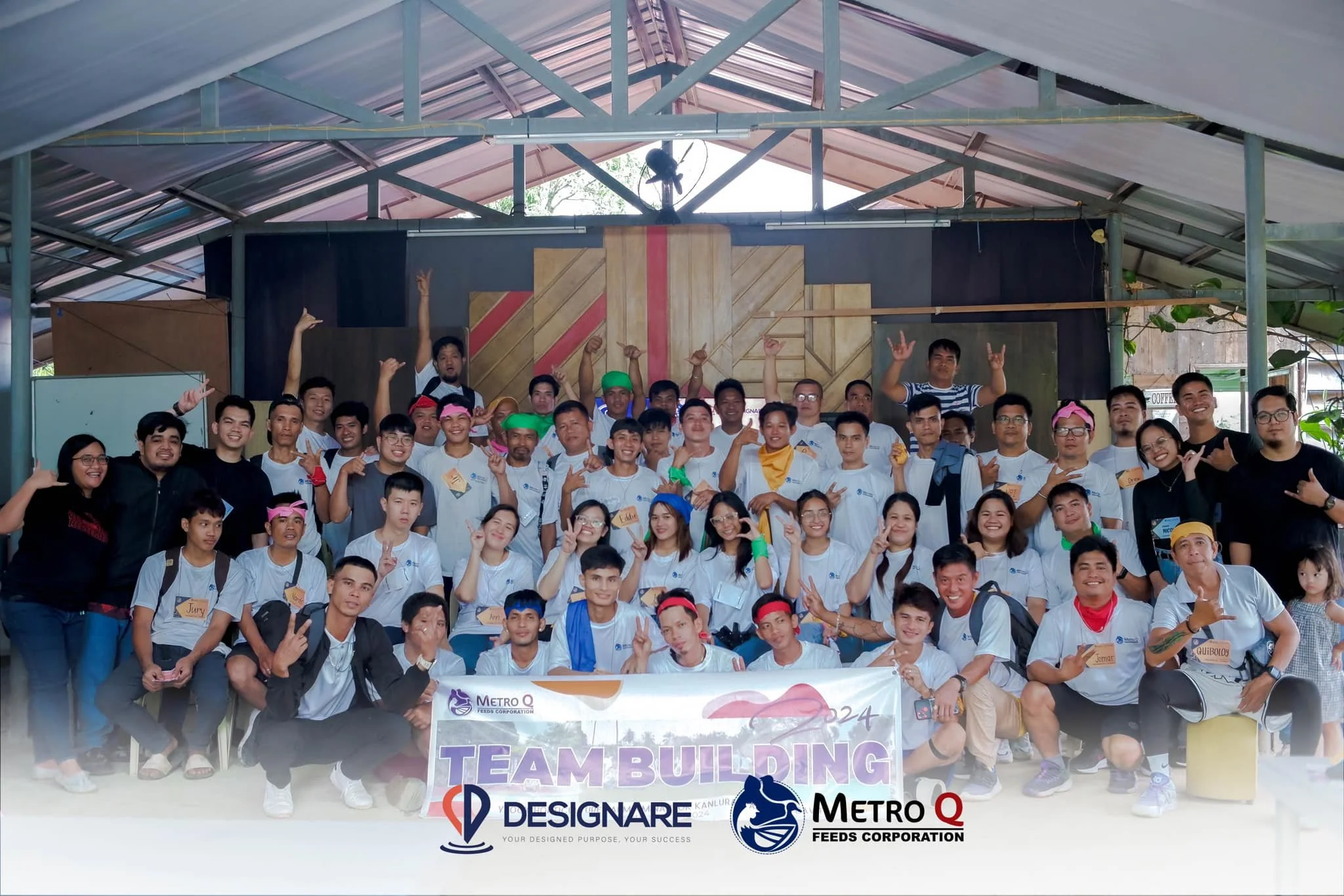 Group of young people and adults posing together indoors for a team-building photo, some making hand gestures, with a banner that reads 'TEAM BUILDING' and logos of Metro Q Feeds Corporation and Designare.