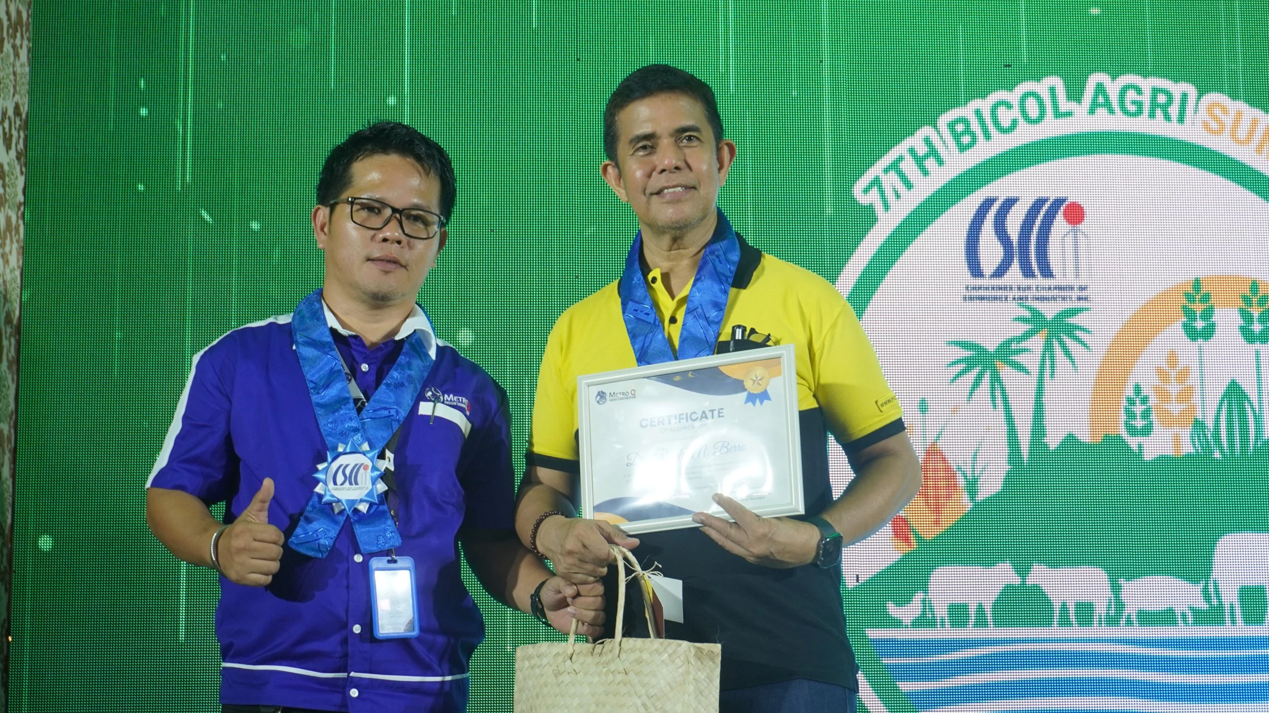 Two men standing on stage at an event, one holding a certificate and a gift bag, both wearing medals around their necks. The man on the left is wearing a purple and white shirt with glasses, and the man on the right is wearing a yellow and black shir
