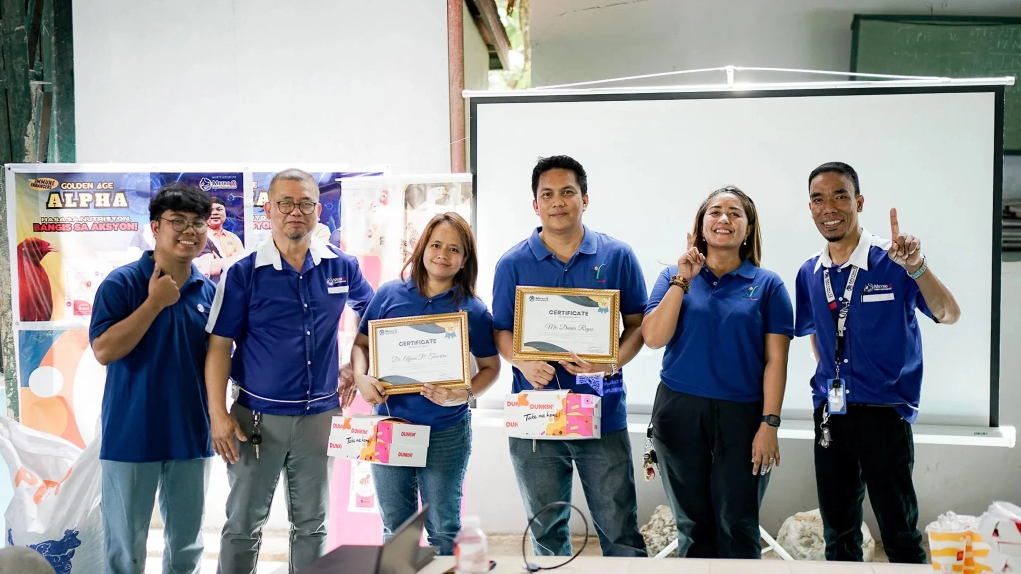 Group of six people standing indoors, smiling, and holding certificate awards, with a banner and whiteboard in the background.