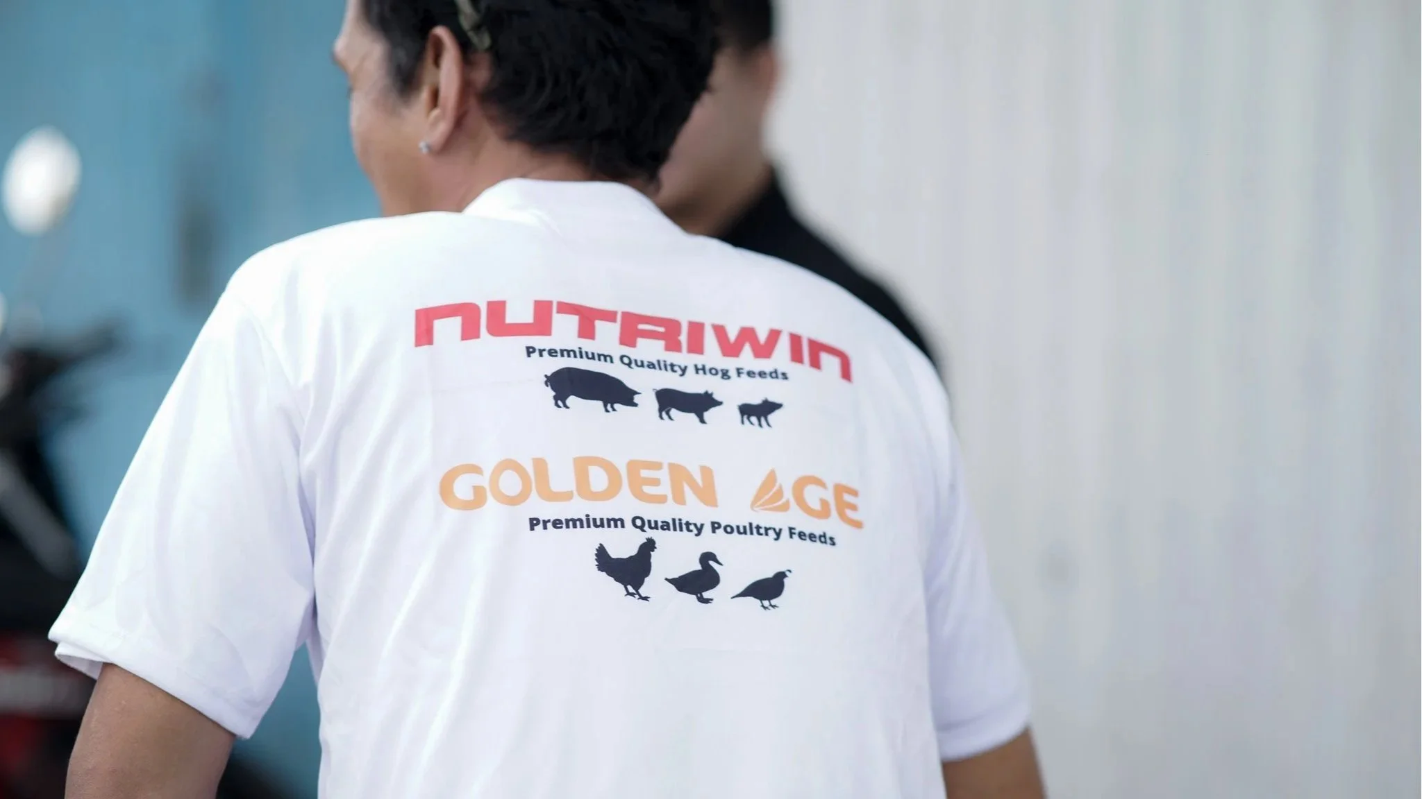 Back of person wearing white shirt with logos and text for Nutriwin and Golden Age, featuring silhouettes of pigs and poultry, advertising animal feed brands.