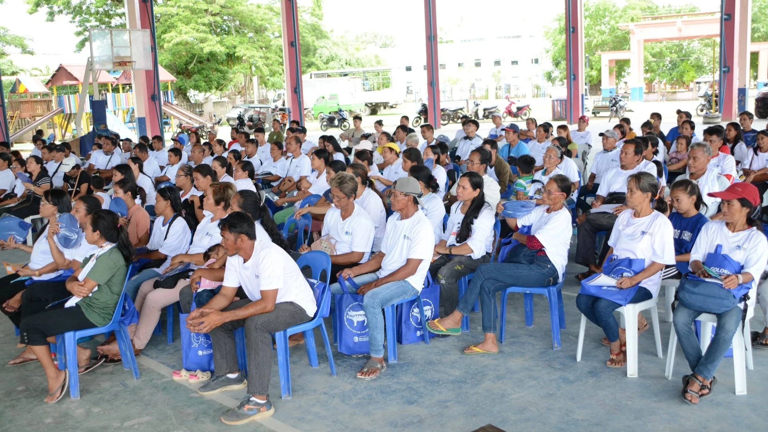 A large group of people sitting on blue and white plastic chairs under a covered outdoor area, attending an event. Some are holding bags, and a few children are present. In the background, there are trees, a playground, and parked motorcycles.