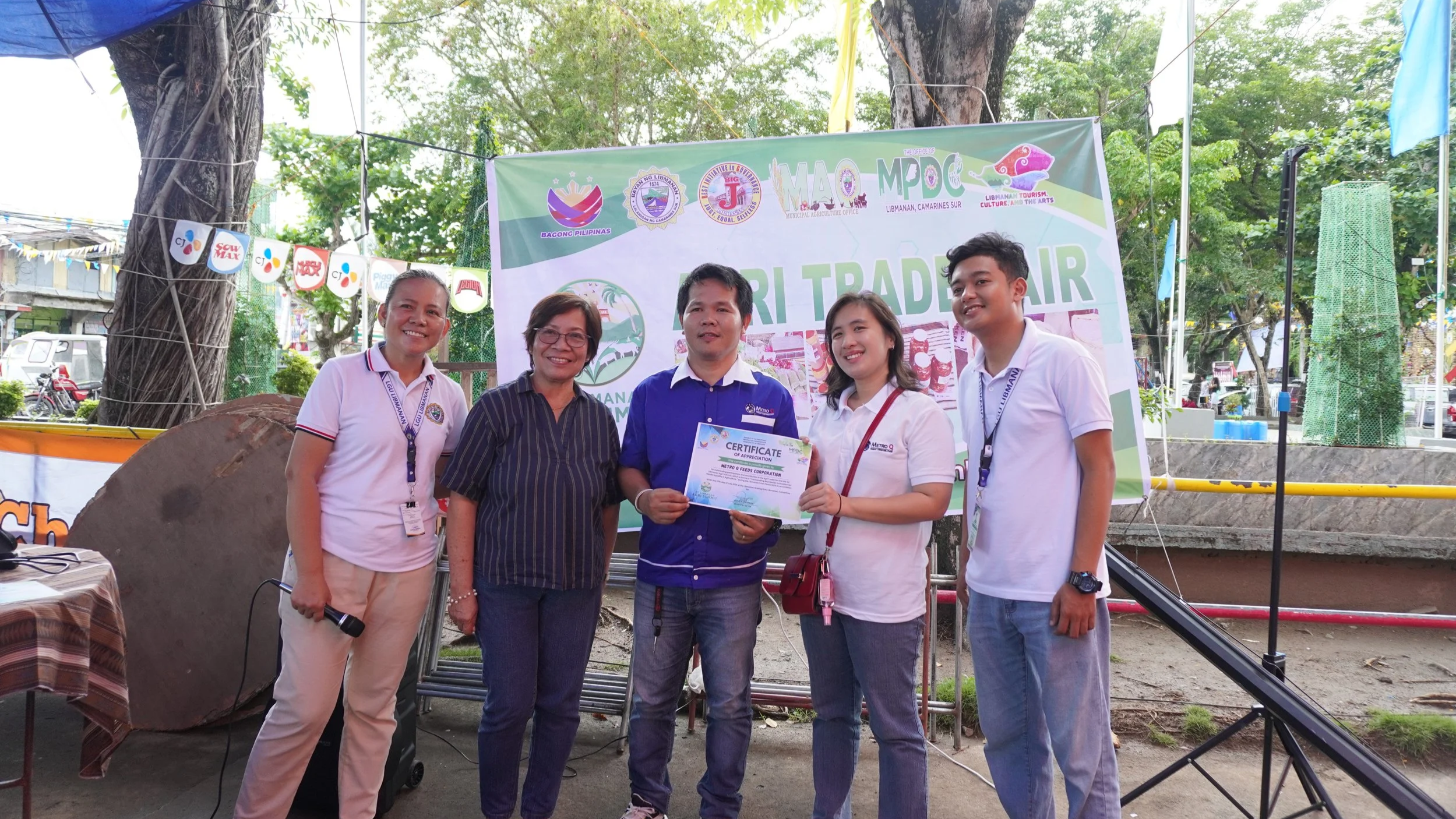 Group of five people standing outdoors in front of a green and white banner, with three women and two men, one man holding a certificate.