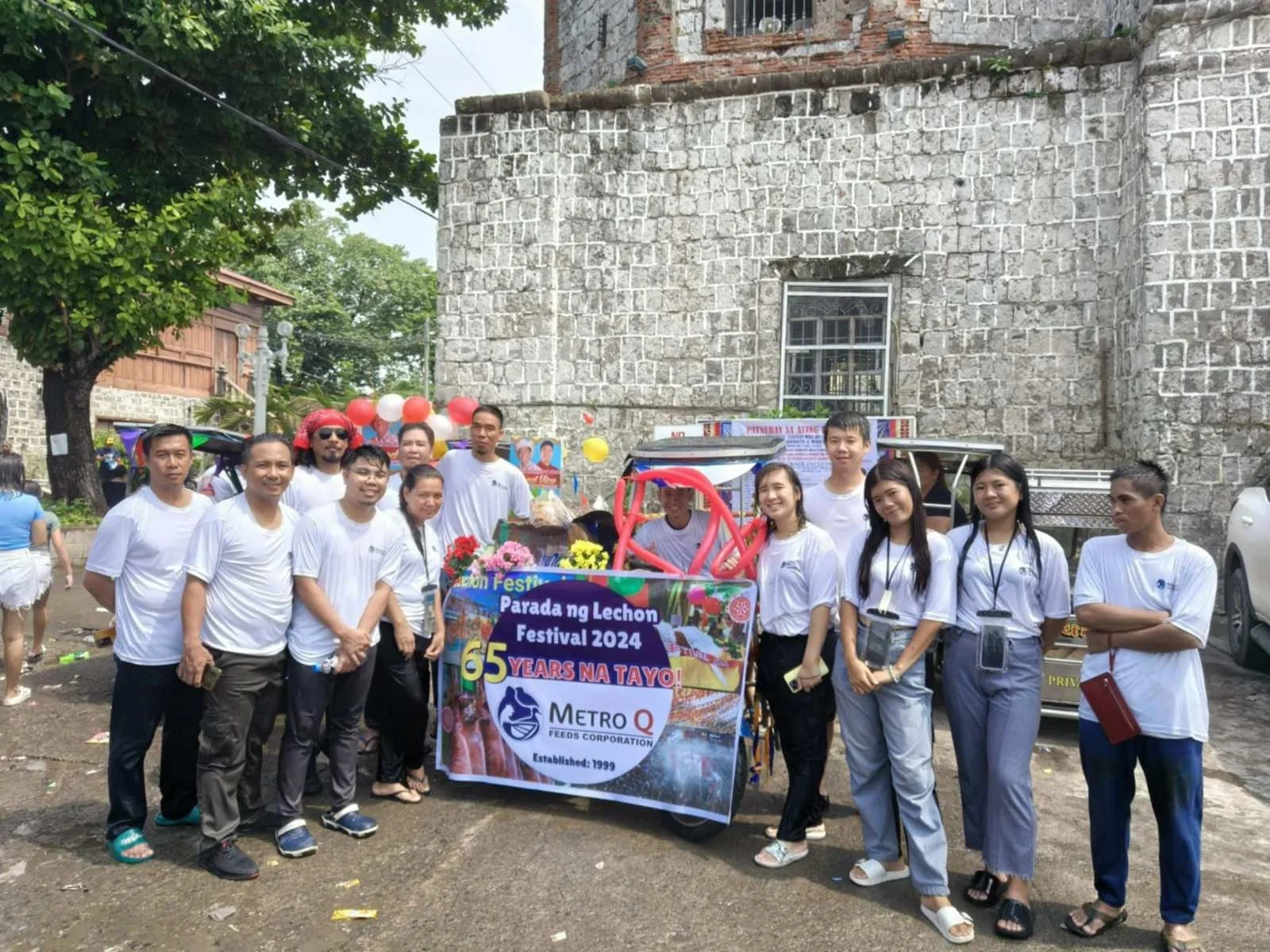 Group of people wearing white T-shirts standing around decorated vehicle for a festival celebration in an outdoor setting, with old stone building and trees in the background.