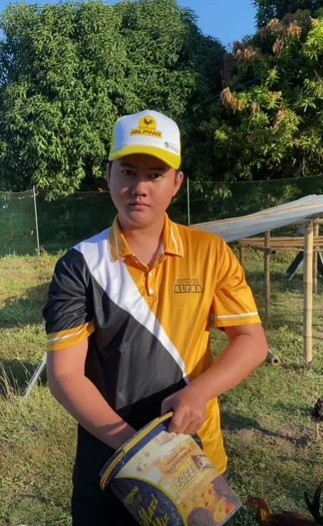 A young boy wearing a white and yellow baseball cap and a yellow, black, and white polo shirt with a logo, standing outdoors with trees and a wooden structure in the background, holding a booklet.