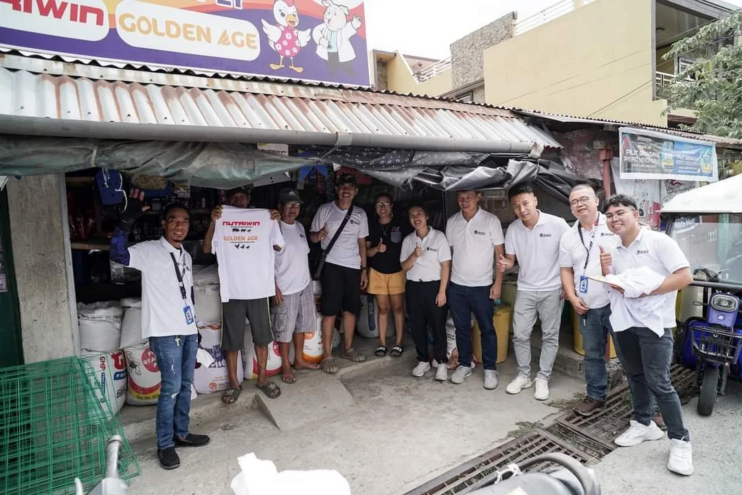 Group of people standing outside a small shop, some in white shirts, smiling and giving thumbs up.