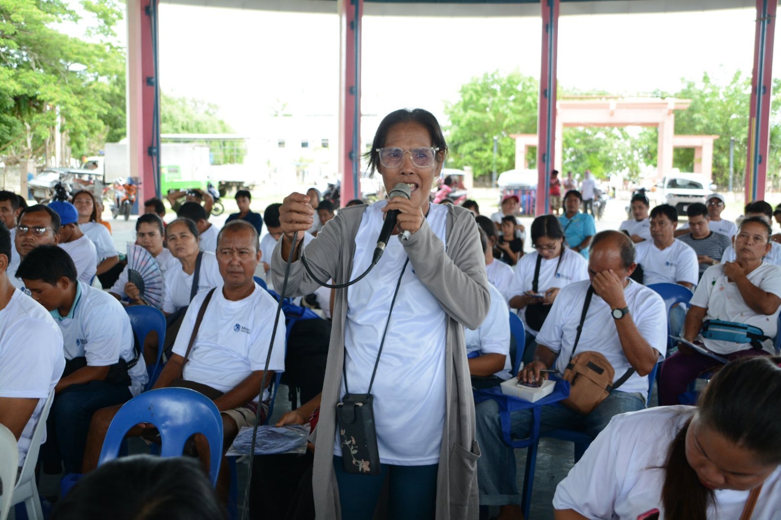 A woman wearing glasses and holding a microphone is speaking to an audience assembled in an outdoor pavilion. The audience is seated in blue plastic chairs, wearing white clothing, with some holding fans or taking notes. Bright green trees and a pink