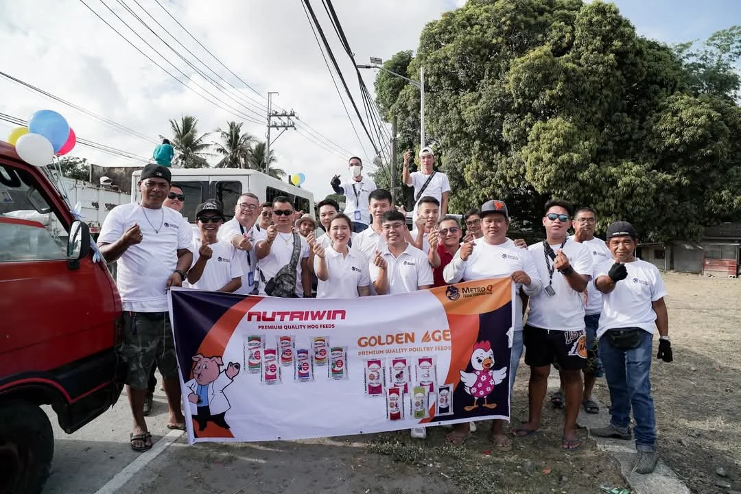 Group of people holding a banner at an outdoor event, with some people giving thumbs up, a red vehicle, trees, electricity poles, and balloons in the background.