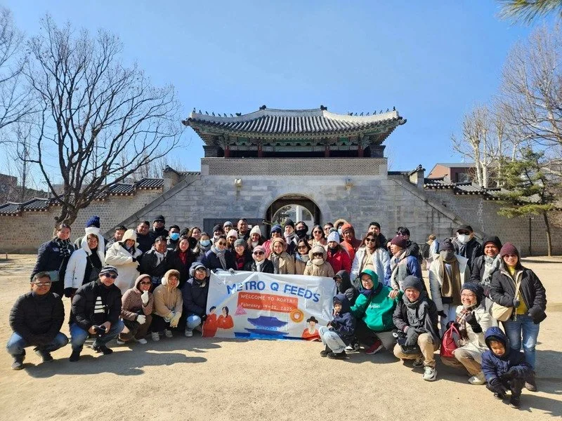 Group of people posing in front of a traditional Asian gate holding a banner that says 'Metro Q Feeds' and 'Welcome to Korea,' on a sunny day.