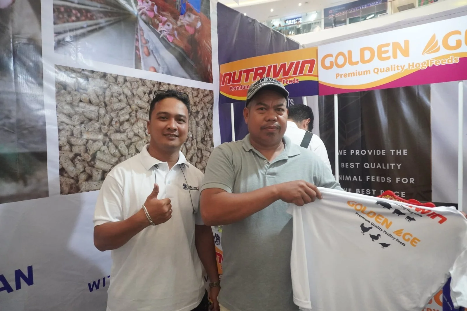 Two men standing in front of a display booth at an event, holding a white bag with logo and images of poultry. One man is smiling and giving a thumbs-up, the other has a neutral expression.