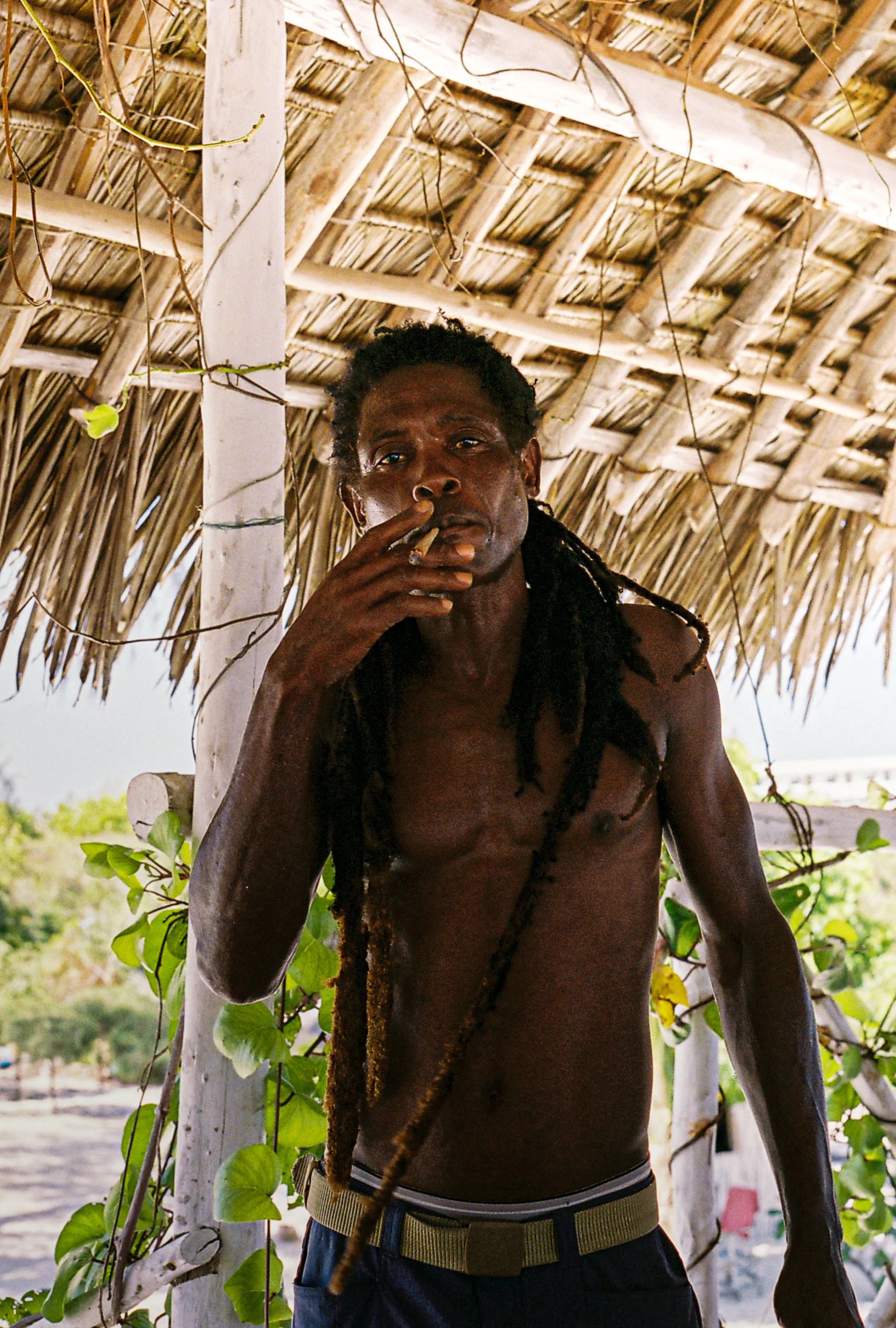 A shirtless man with dreadlocks standing under a thatched roof structure, smoking a cigarette, with green leafy vines around him and a blurred outdoor background.