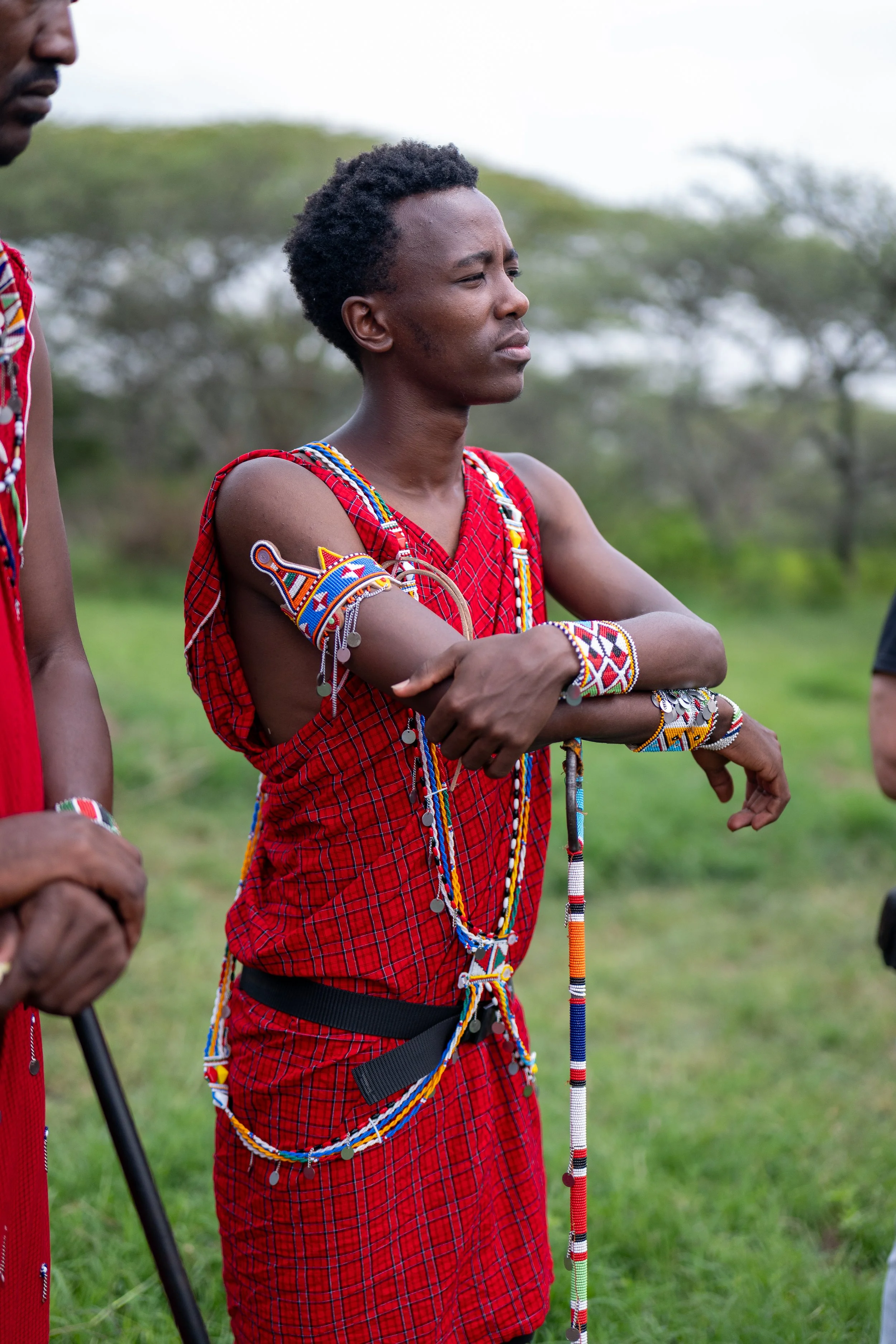 A young woman wearing traditional Maasai attire, including a red checked shuka and multiple beaded jewelry, standing outdoors with arms crossed, during daytime with a background of trees and a cloudy sky.