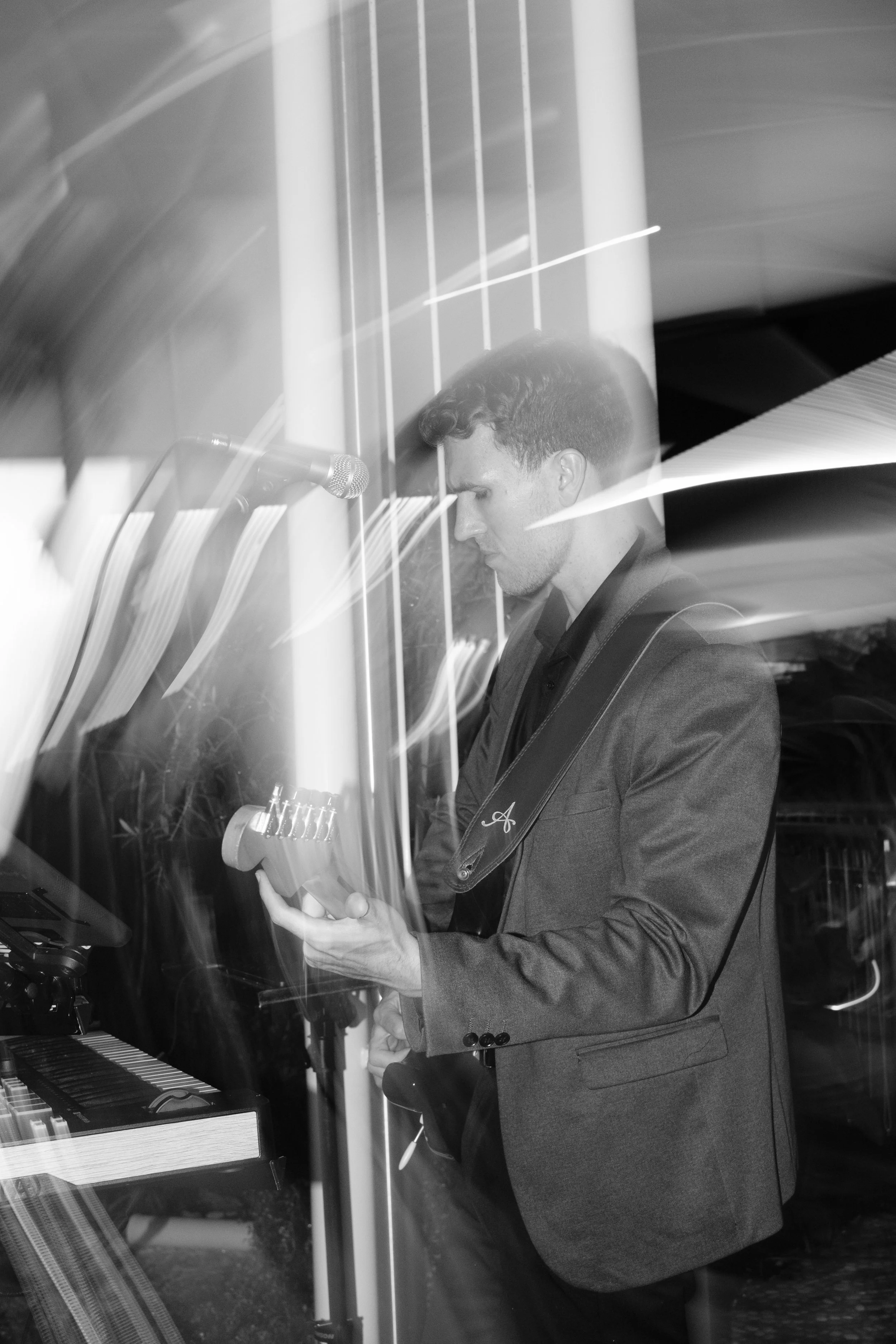 A man in a gray suit playing a black grand piano, holding a sheet of music, with a microphone nearby, indoors with modern lighting and reflective surfaces.