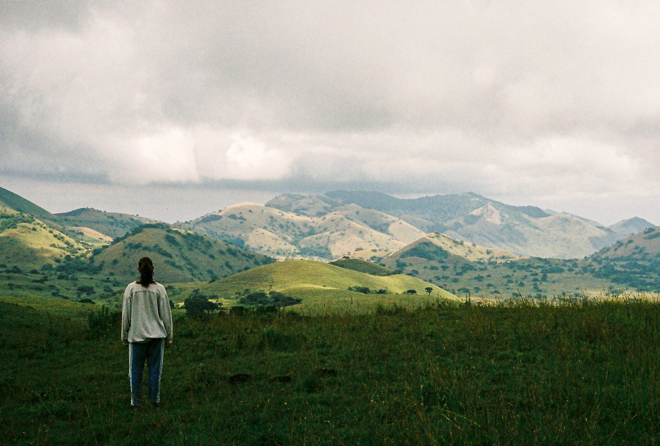 A person with long hair wearing a loose shirt and blue jeans standing alone in a green grassy area, facing away, overlooking rolling hills and mountains under a cloudy sky.