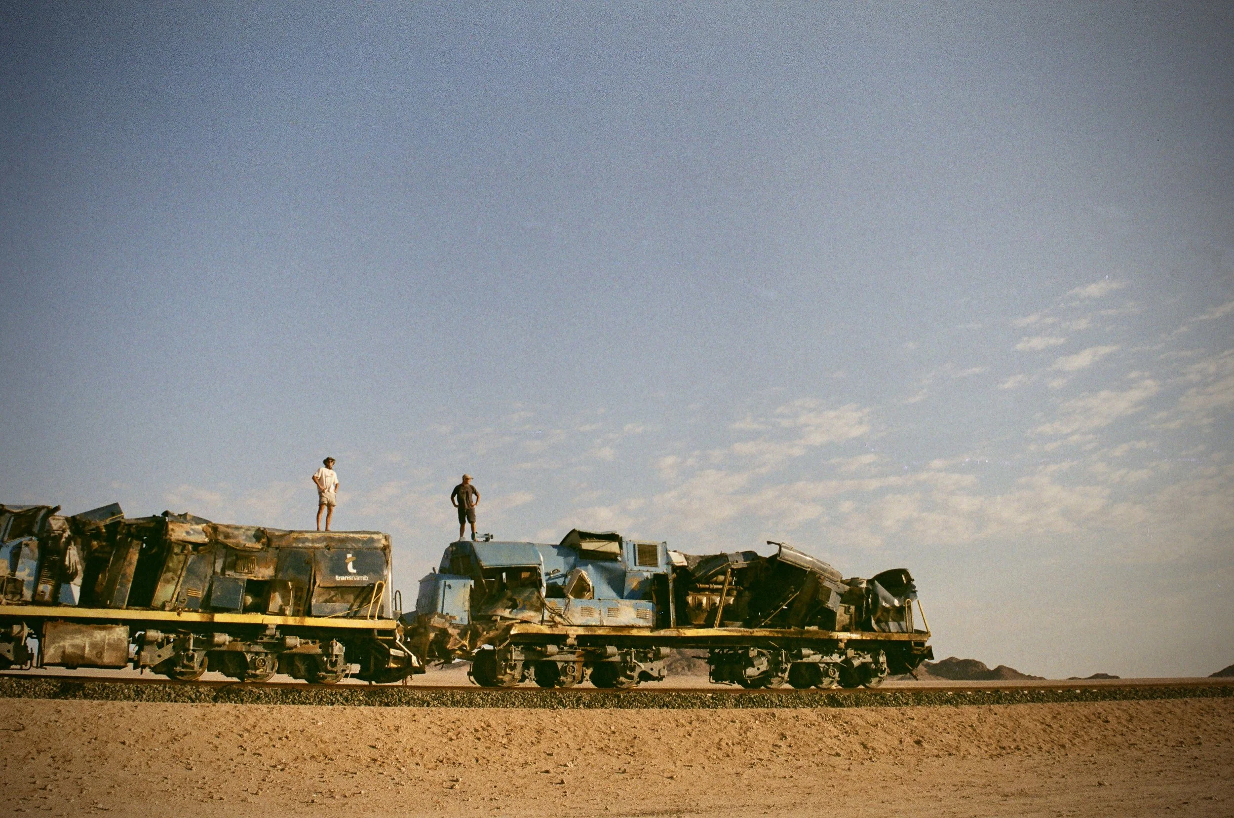 Three people standing on top of a large, rusty, disassembled train car in a barren desert landscape under a mostly clear blue sky.