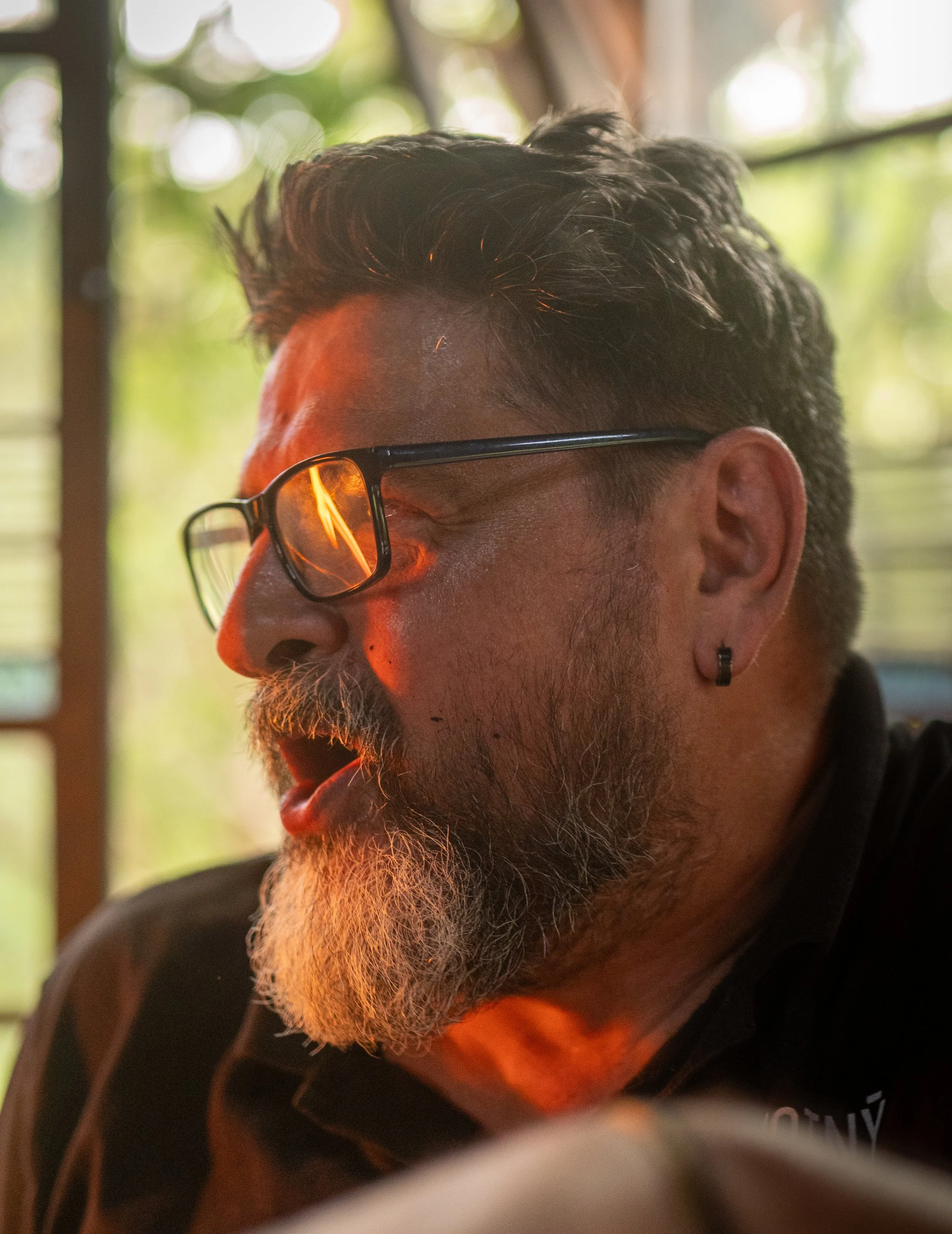 Close-up of a man with glasses and a beard, wearing a black shirt with orange accents, outdoors with greenery and windows in the background.