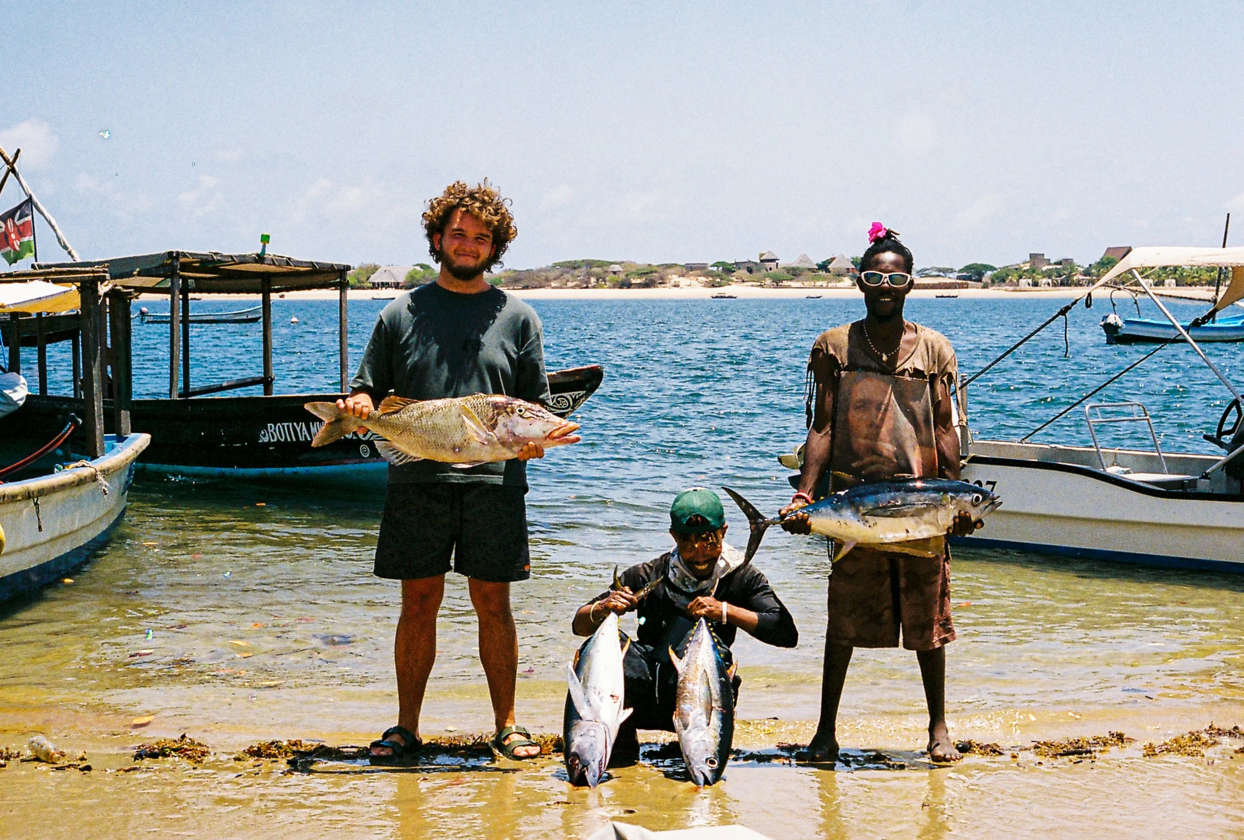 People at the shoreline displaying fish they caught during a fishing excursion, with boats in the water and a distant shoreline with buildings and trees.