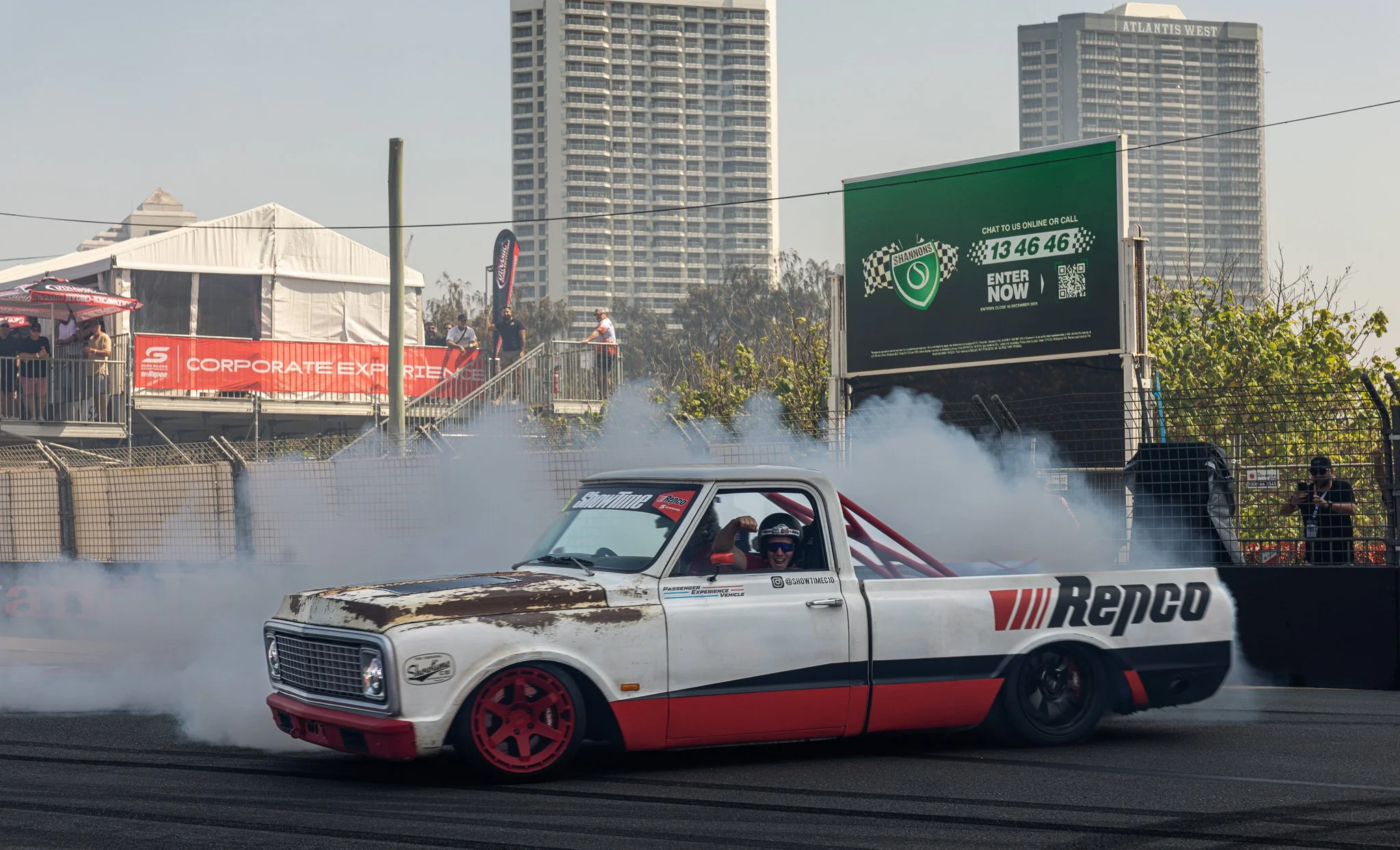 A race car with Repcos branding drifting on a track with tire smoke, a driver smiling and waving, background includes a viewing platform with people, banners, and high-rise buildings.