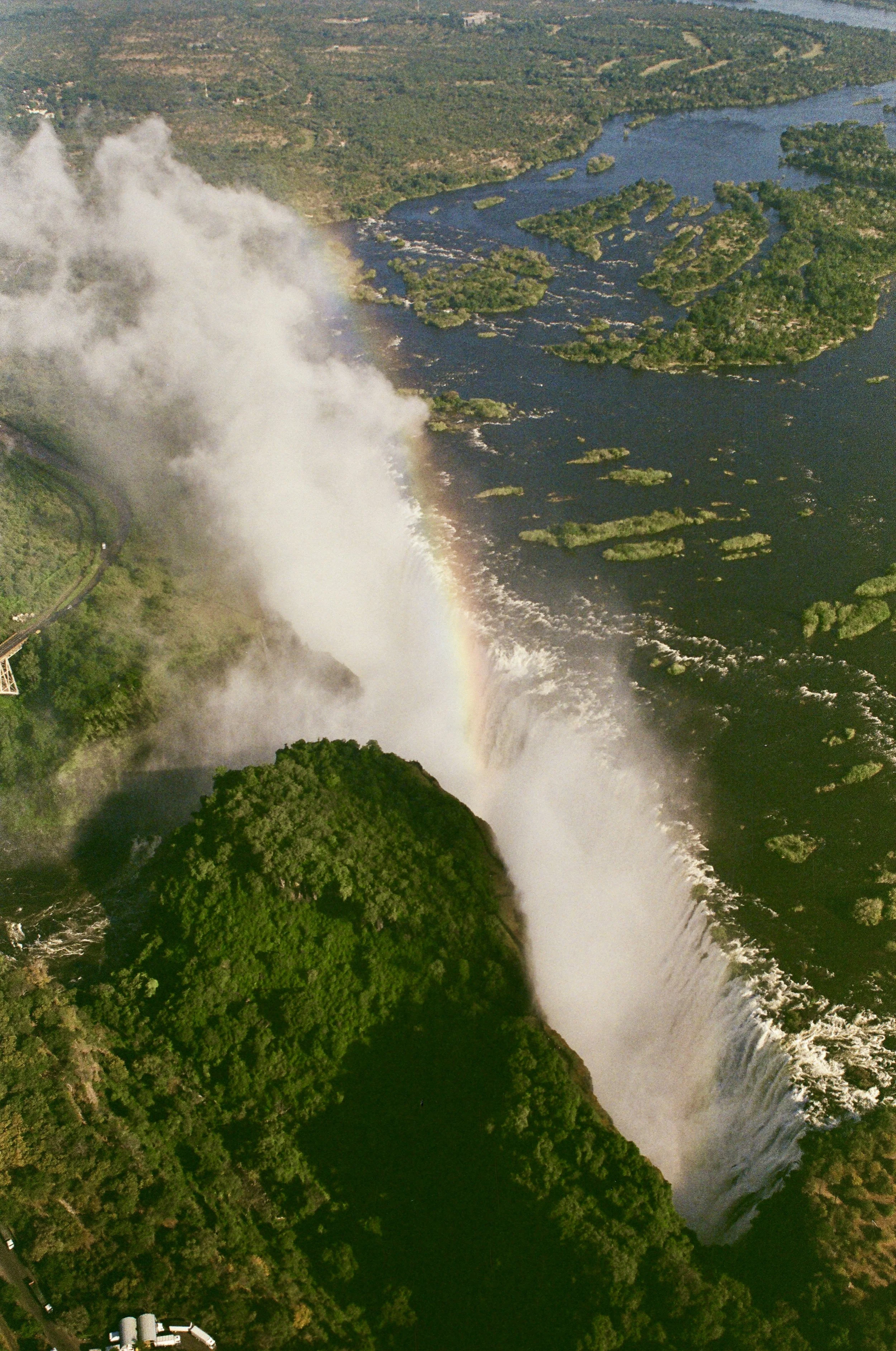 Aerial view of a large waterfall with a rainbow forming in the mist, surrounded by lush green trees and water bodies.