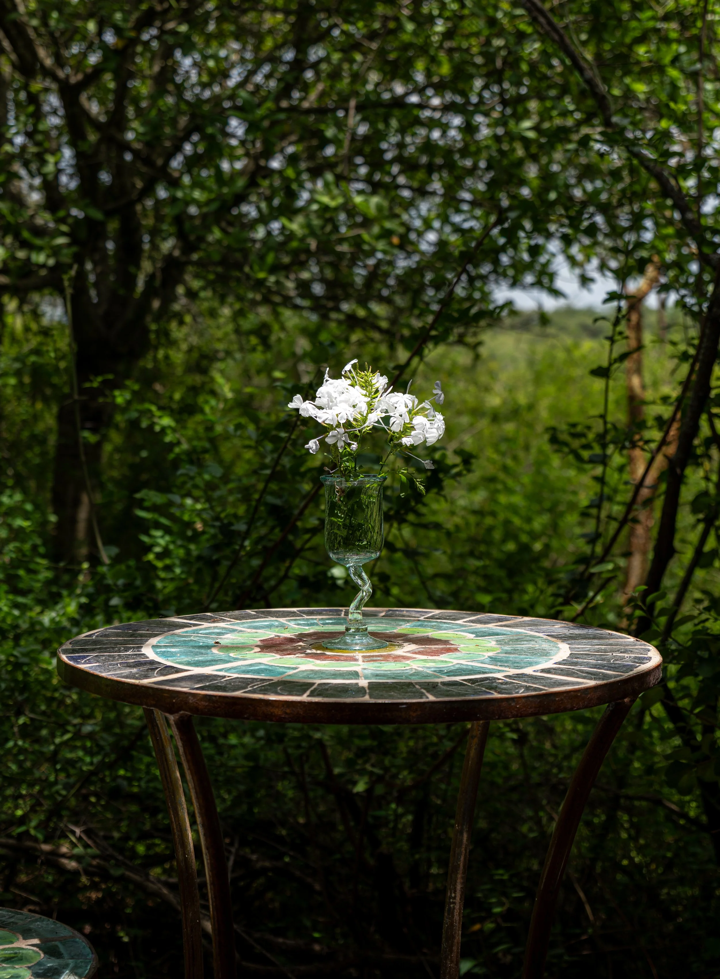 A white flower arrangement in a tall, intricately shaped green glass vase on a mosaic-tiled table outdoors, surrounded by lush green foliage.