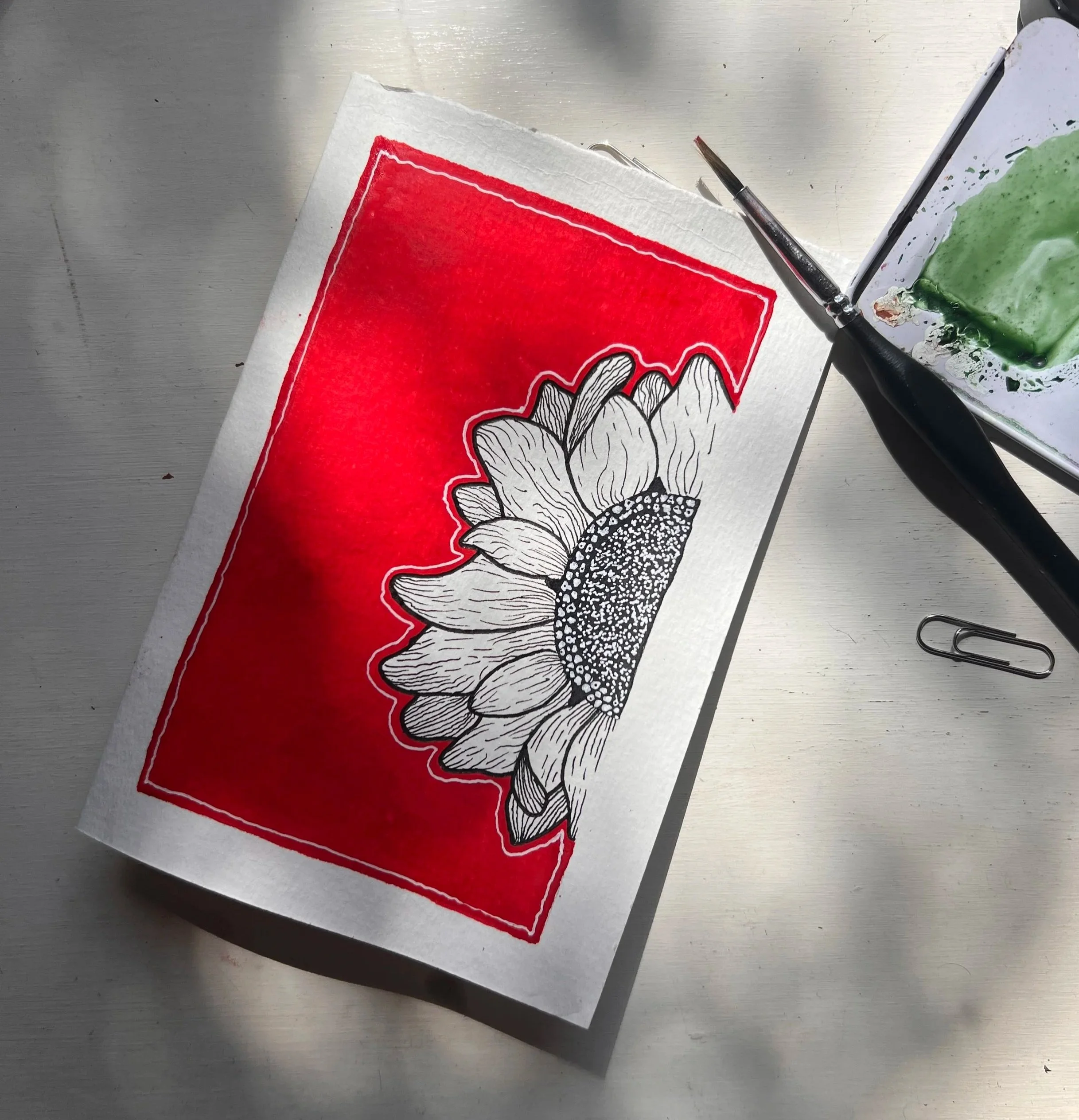 A piece of paper with a colorful flower drawing and a background of a red rectangle, placed on a metal surface alongside a paintbrush and part of a palette with green paint.