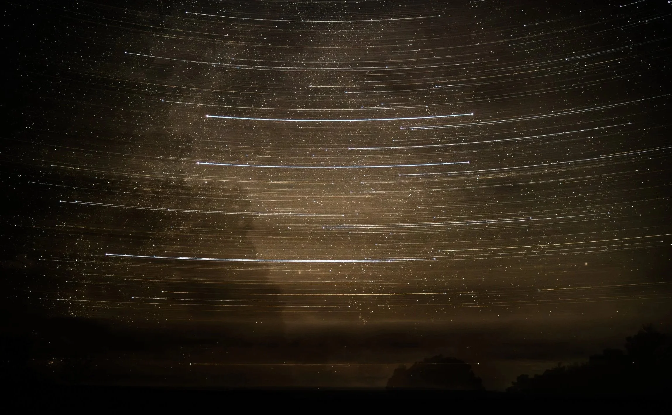 Long exposure photograph of night sky with star trails and stars visible over dark landscape.