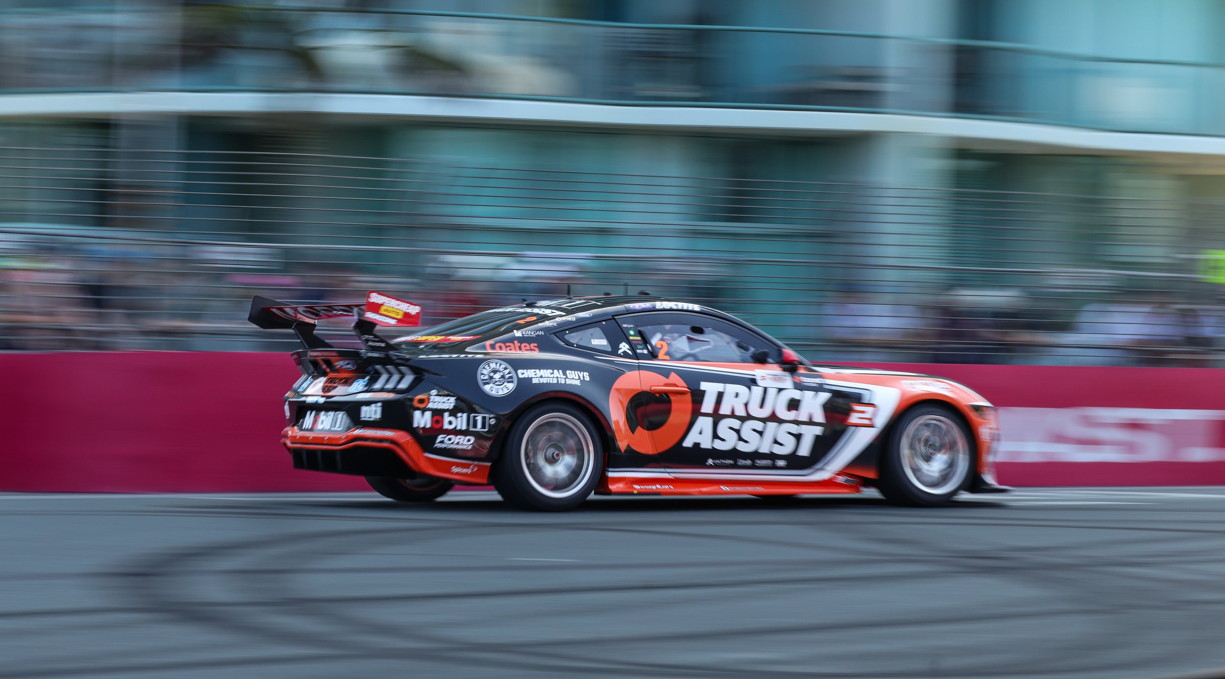 A race car with a black, orange, and white livery speeding on a race track, with a blurred background of a glass building and safety barrier.