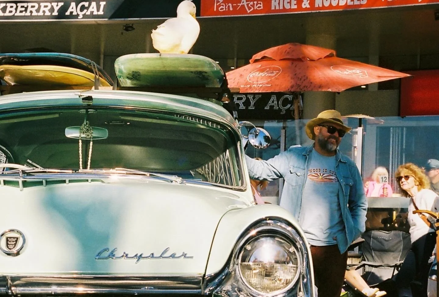 A vintage white Chrysler car with roof-mounted surfboards at an outdoor event. A man in a denim jacket, sunglasses, and a wide-brimmed hat stands next to the car, smiling. Several women are in the background.