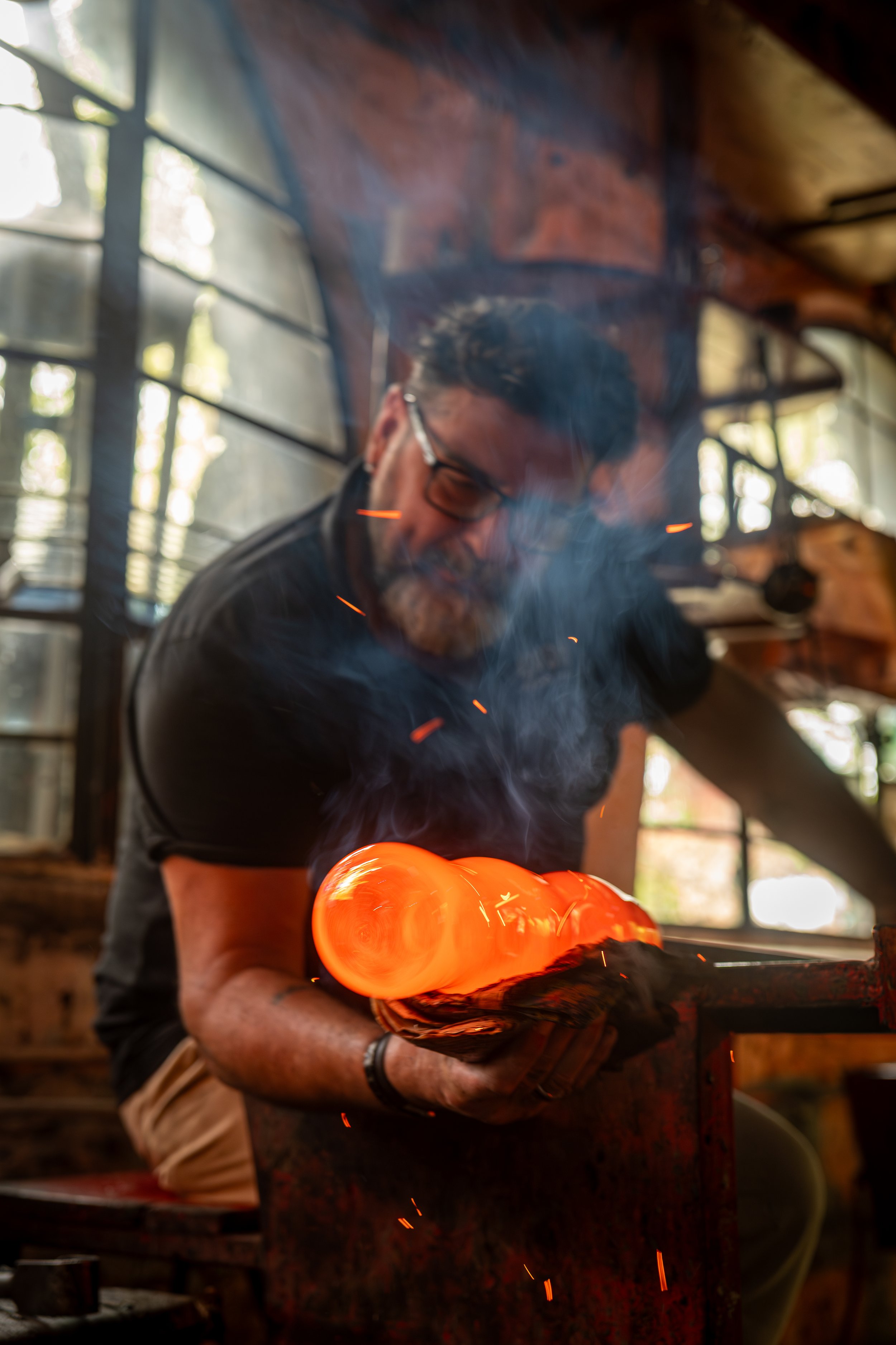A person wearing safety glasses holding a glowing piece of metal in a workshop, with sparks and smoke around.