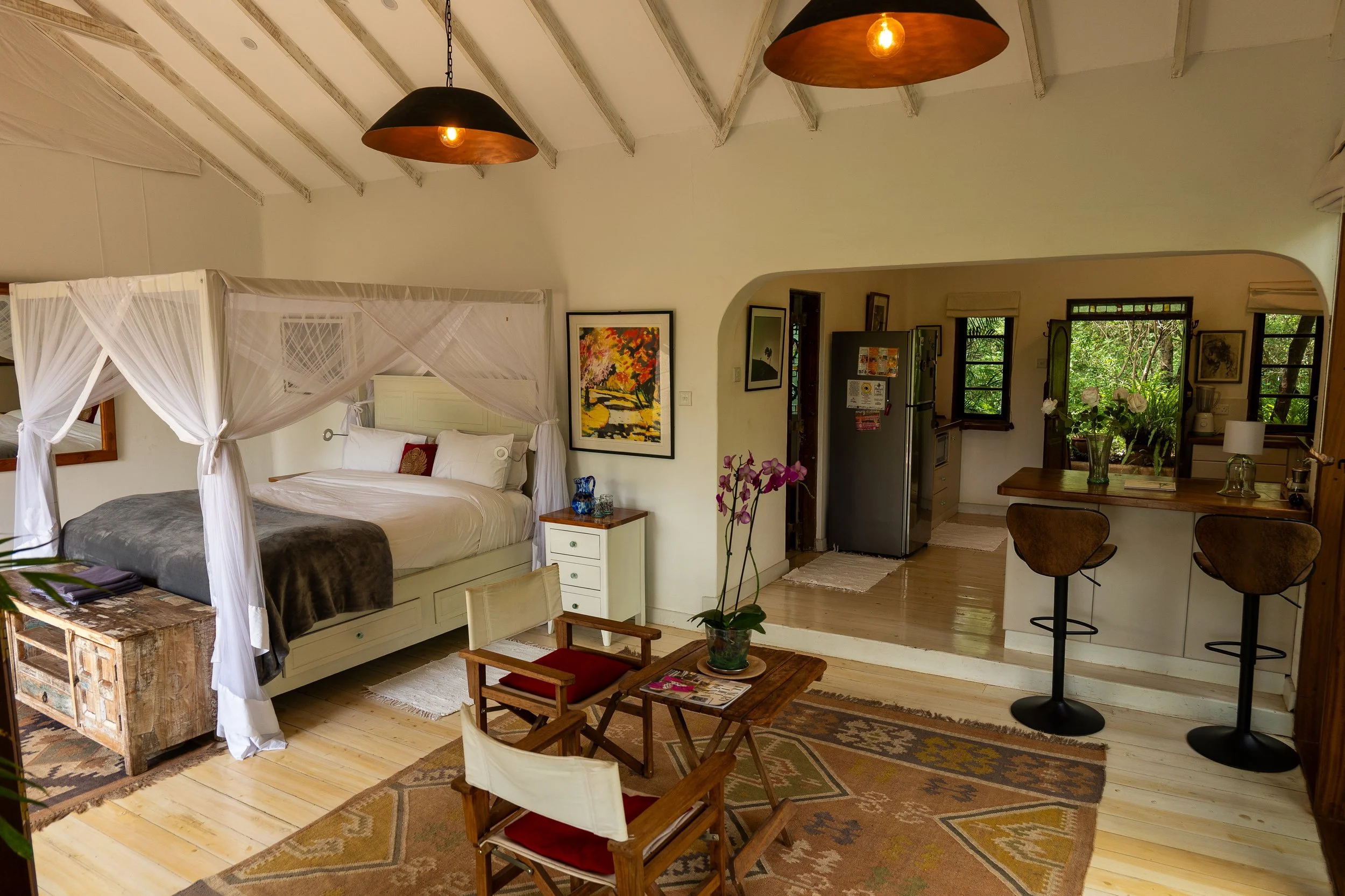 A cozy living room with a four-poster bed, colorful artwork on the walls, a wooden table with a pink orchid, and a kitchen area with a refrigerator and open windows showing greenery outside.