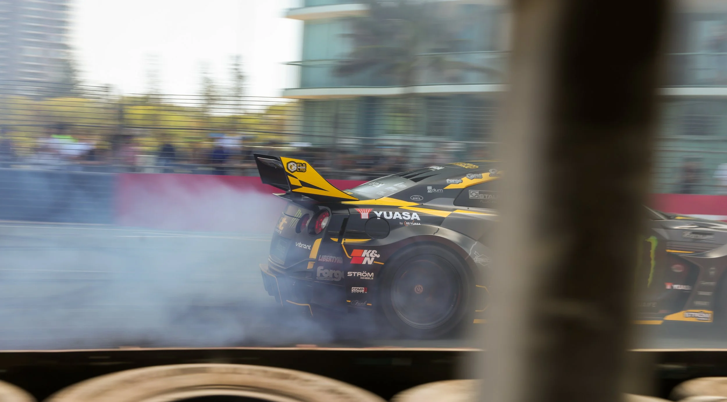 A race car with a black and yellow livery speeds around a track, creating smoke from the tires, and is partially seen through a blurred barrier or screen.