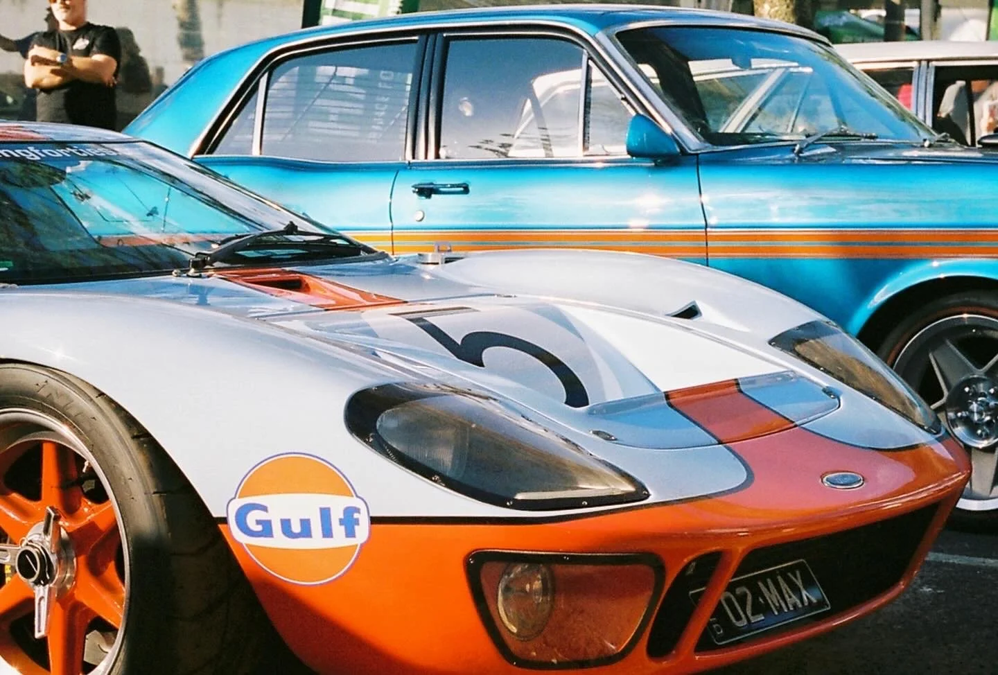 A classic race car with Gulf racing livery, orange, white, and blue, with the number 5 on the hood. Behind it is a vintage blue car with a stripe design.