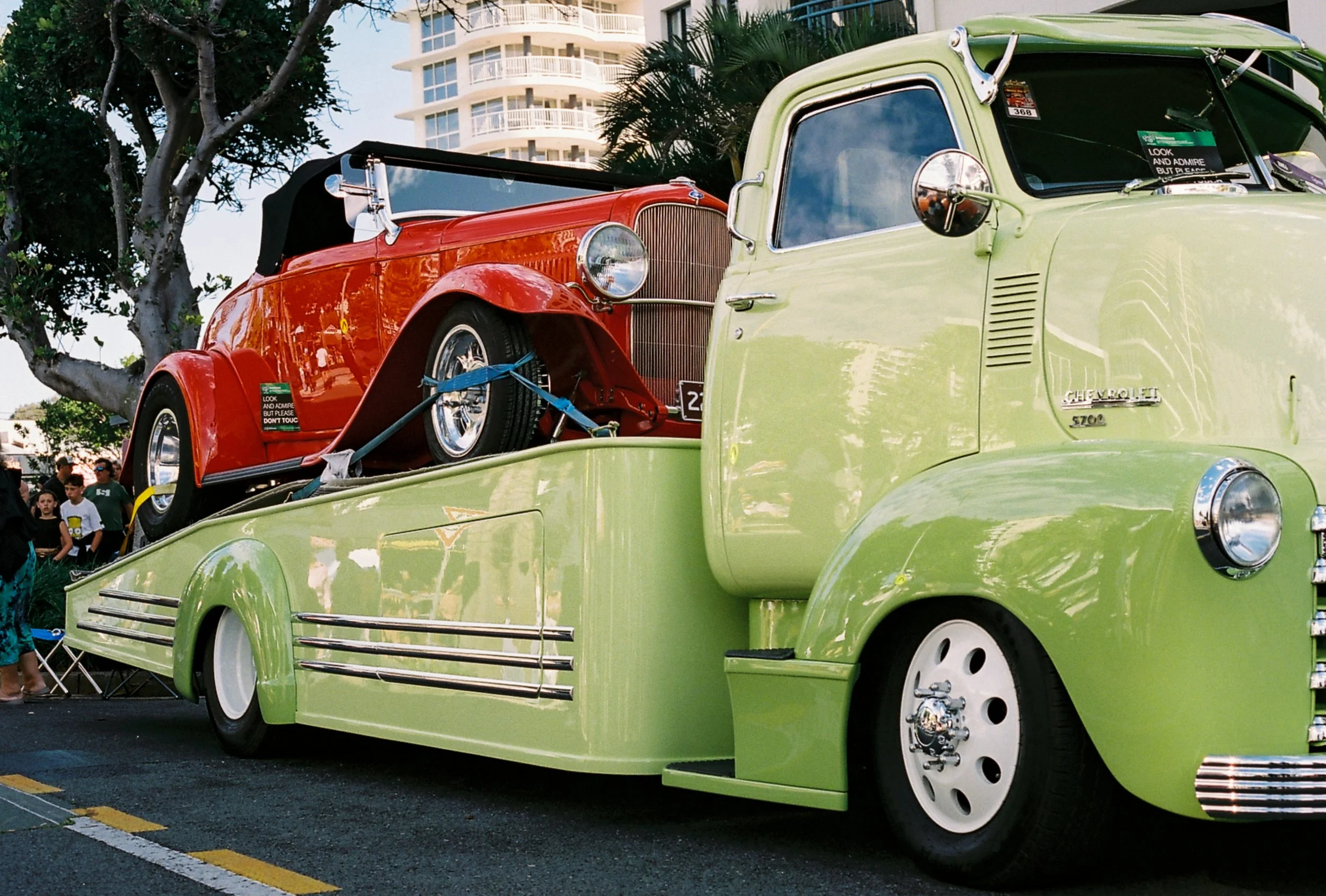 A vintage green Chevrolet truck with a red classic car on its flatbed at an outdoor car show, with people and a modern apartment building in the background.