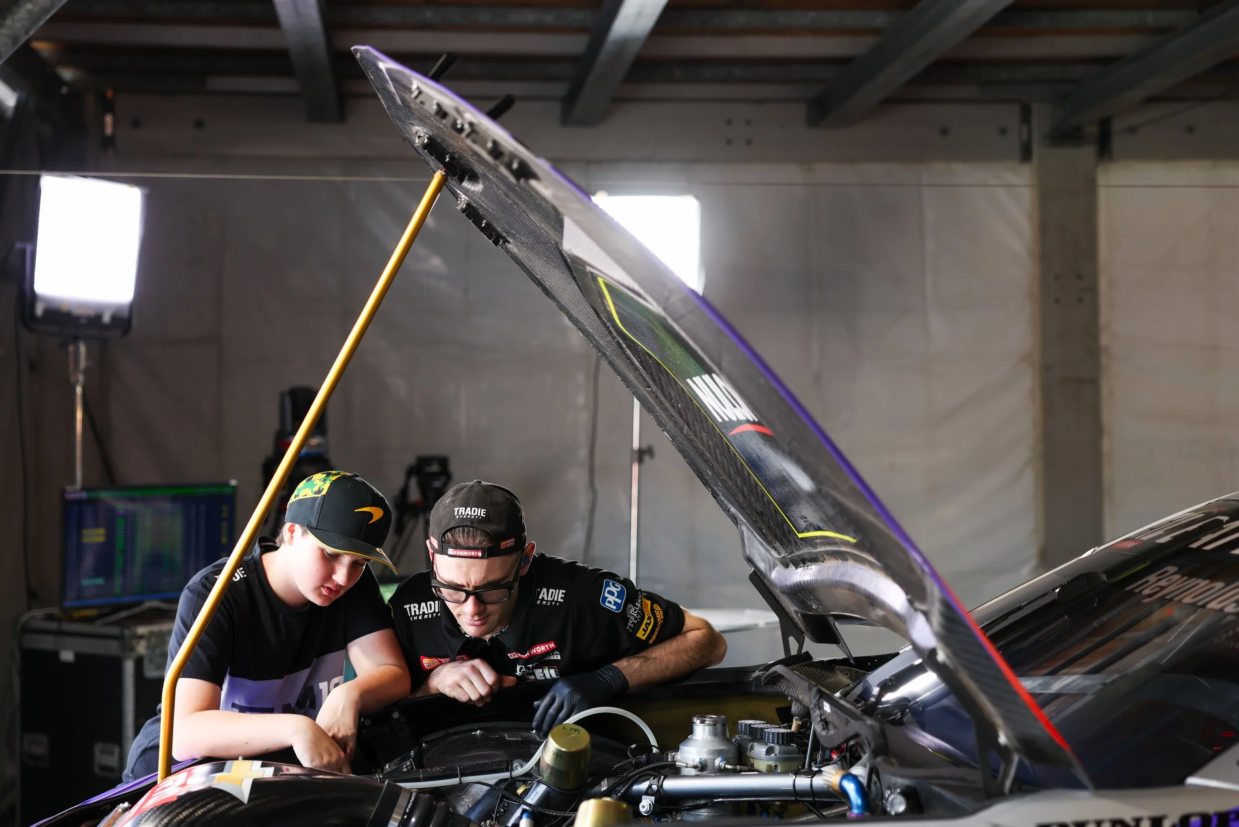 Two mechanics working under the hood of a race car in a garage, with team members and equipment visible in the background.