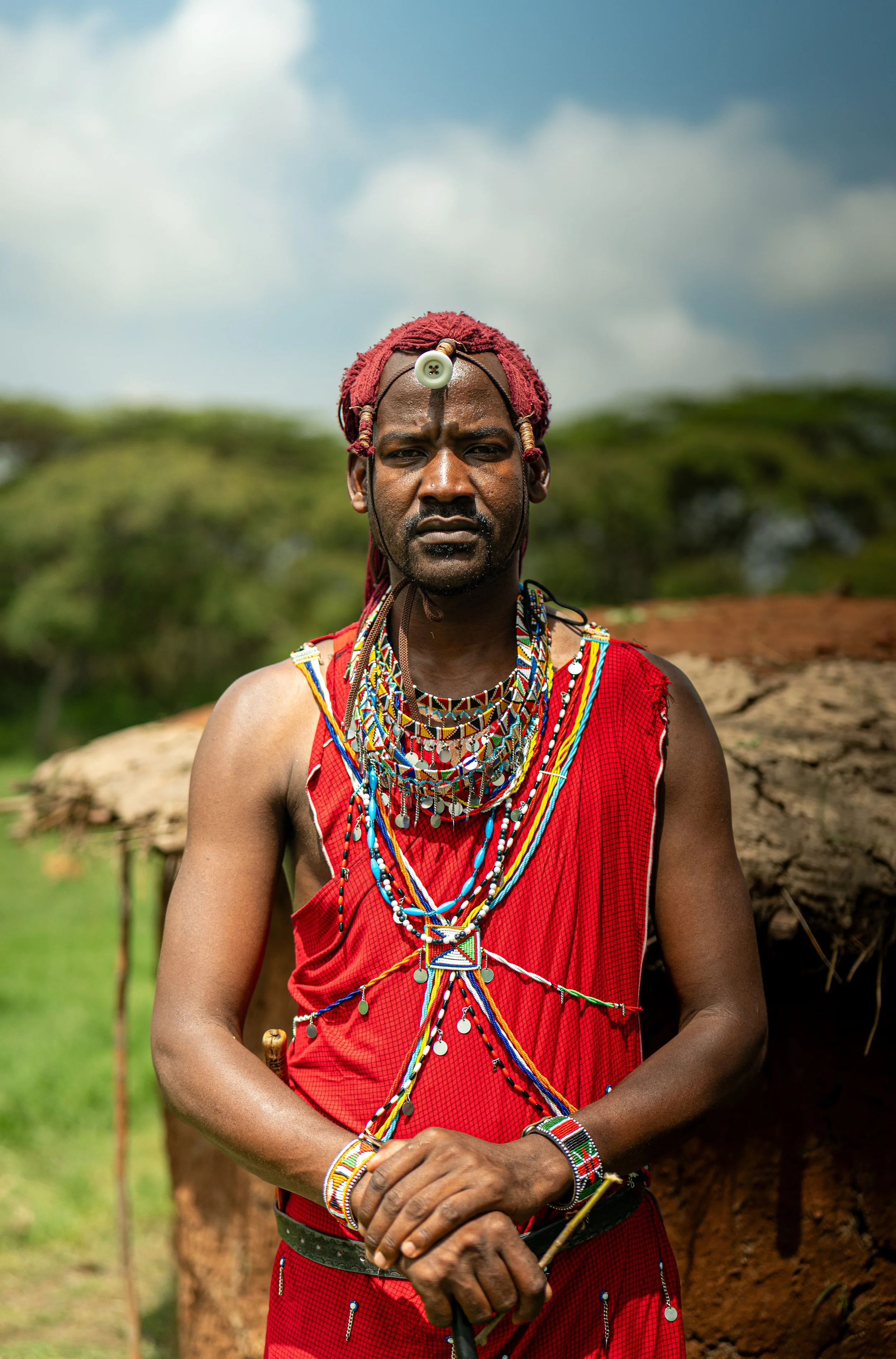 A Maasai man dressed in traditional red attire and jewelry, standing outdoors with a blurred background of trees and sky.