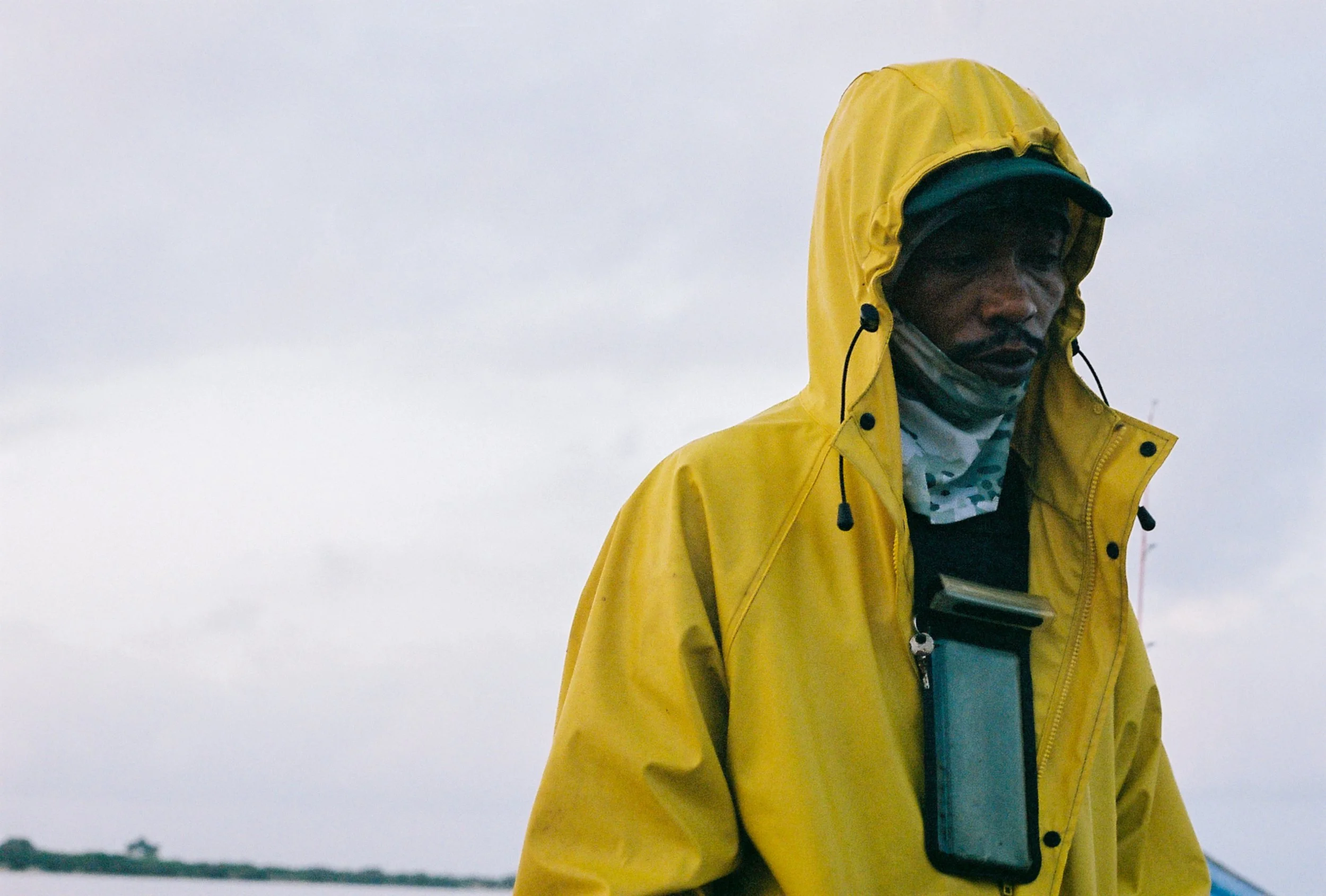 A person wearing a yellow waterproof jacket, a cap, and a face mask, holding a camera or smartphone outdoors in a cloudy, overcast environment.