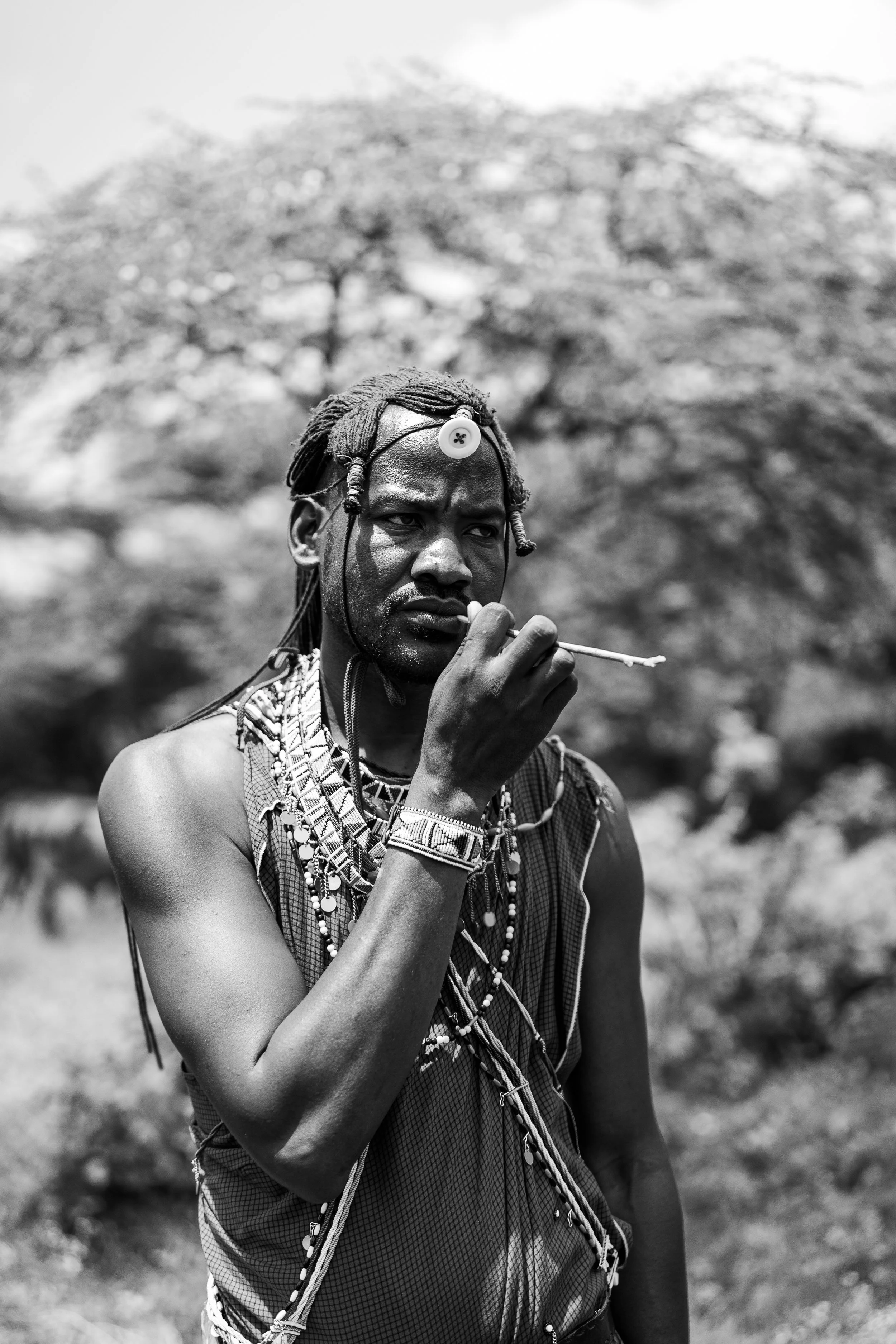 A black and white photo of a man with traditional Maasai attire, wearing beaded jewelry and a headpiece with a button, holding an object near his mouth, outdoors with trees in the background.