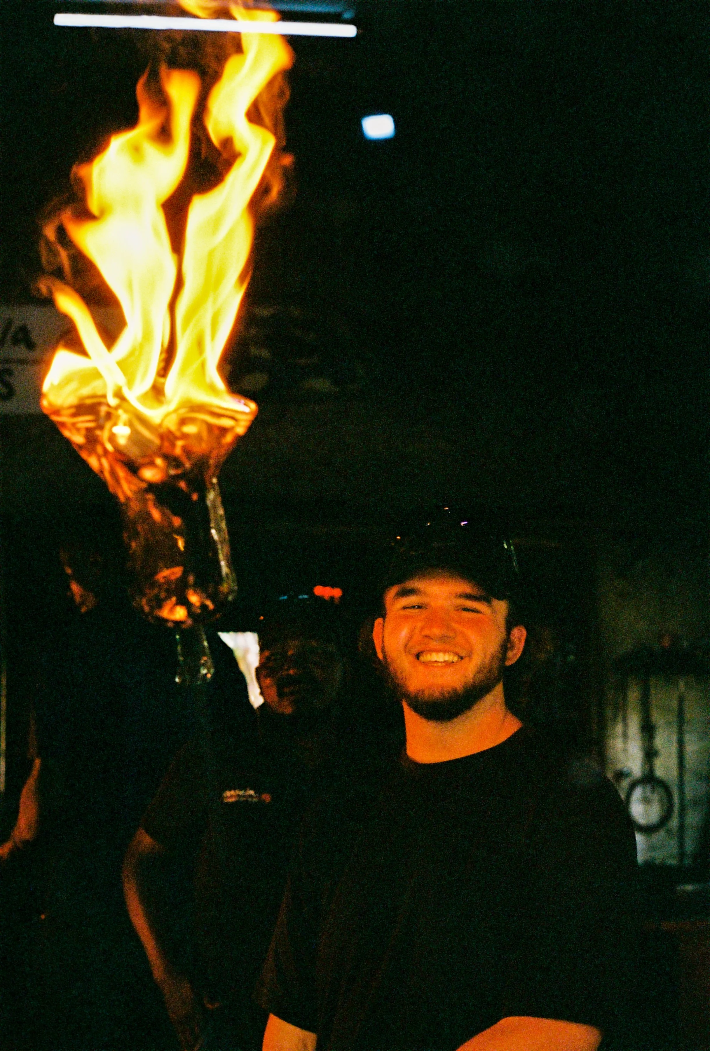 A cheerful young man holding a lit torch with flames at night, smiling at the camera.