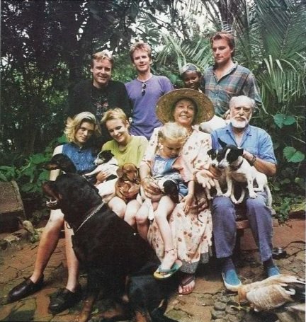 Group of nine people, children and adults, sitting outdoors with dogs and sitting on a bench in a lush, green garden.