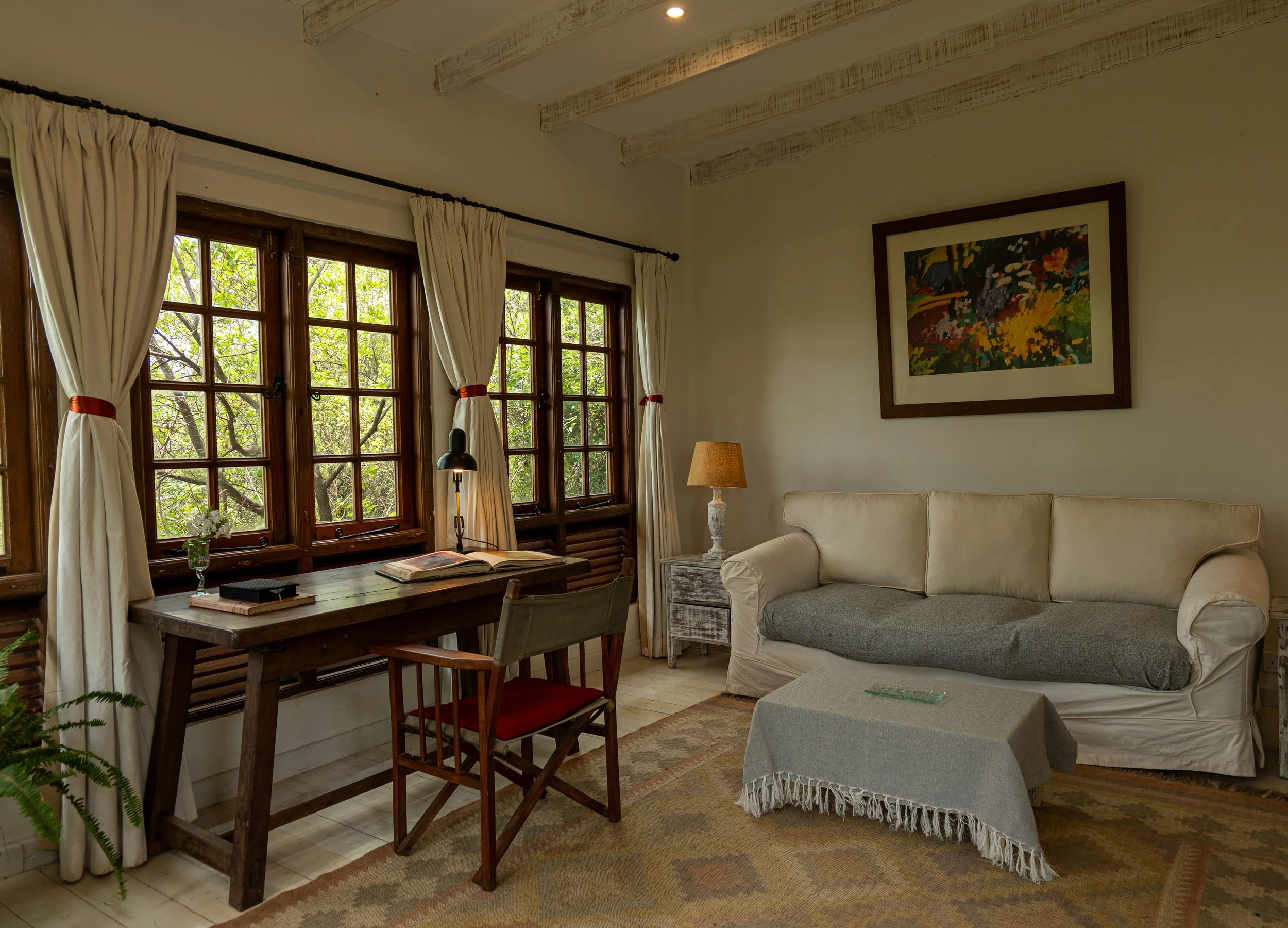 A cozy living room with large windows, white curtains tied back, providing a view of green trees outside. There is a wooden dining table with books and a glass of flowers, surrounded by chairs. A beige sofa with a matching throw and tapestry on the wall adds to the warm decor. A table lamp and a small side table with a lamp are also visible.
