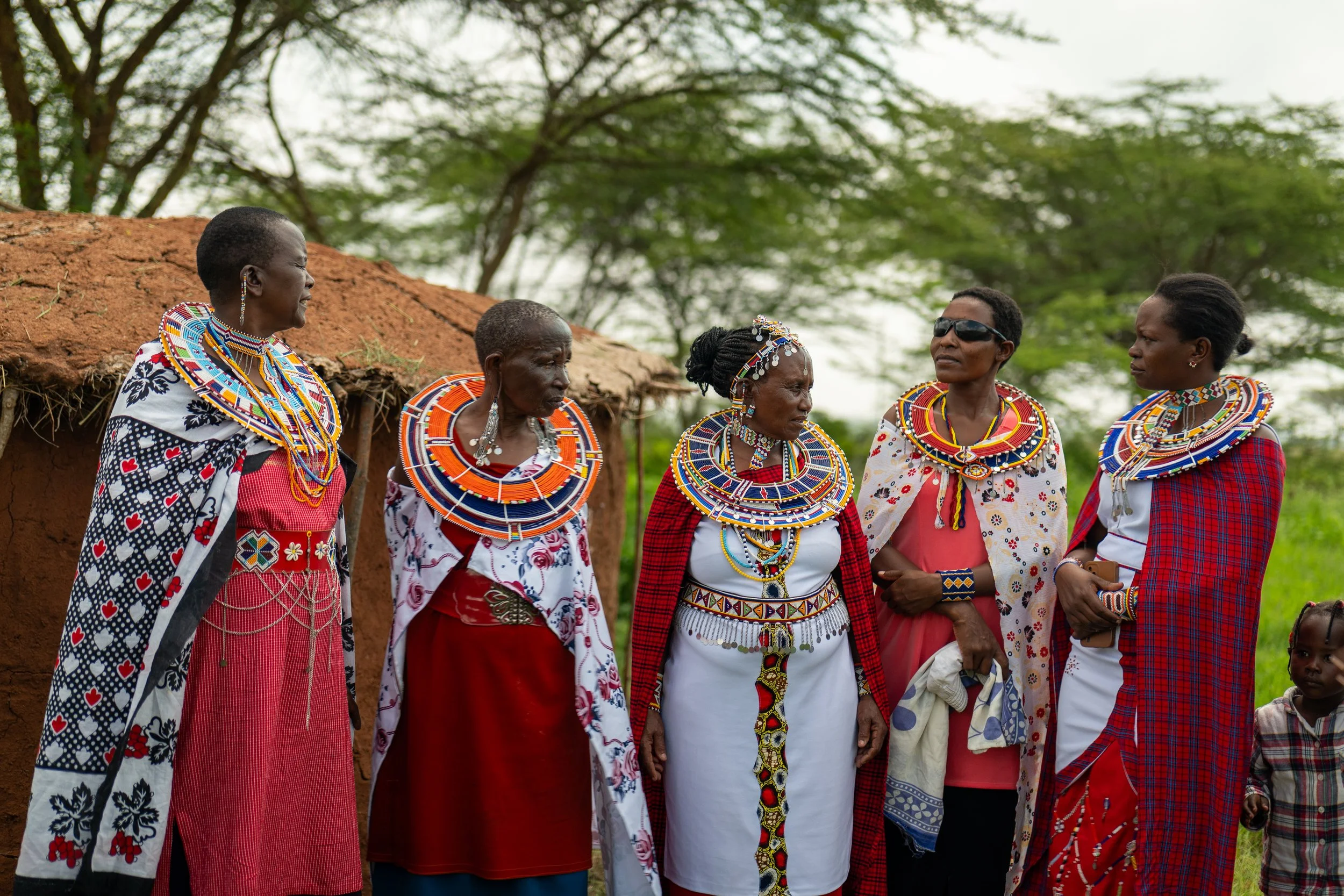 Group of women wearing traditional Maasai beads and clothing standing outdoors near a building with a thatched roof, with trees and greenery in the background