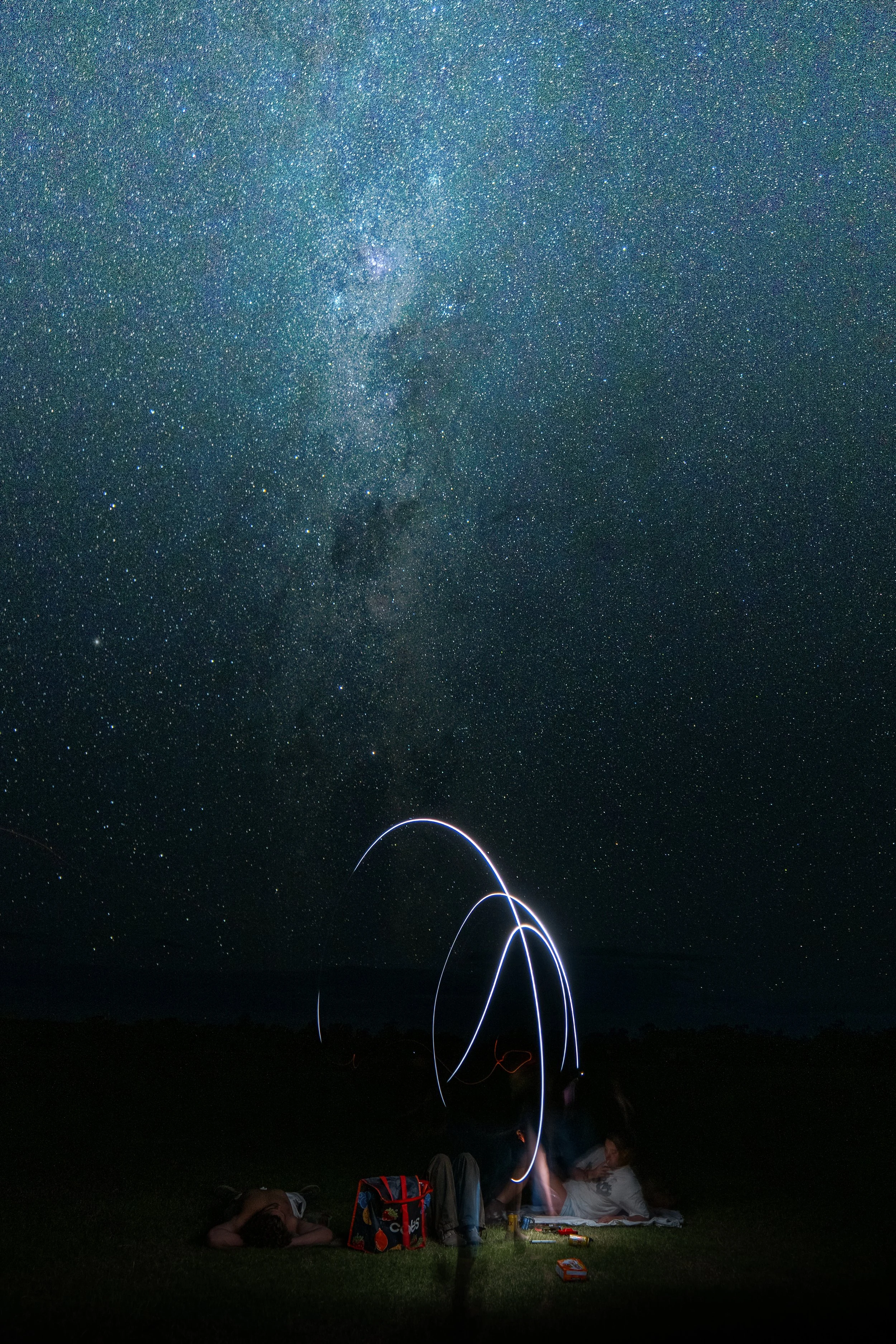 Children sitting on grass at night, observing the starry sky, some lying down, with light trails of fireworks or sparklers in the sky and camping items around them.