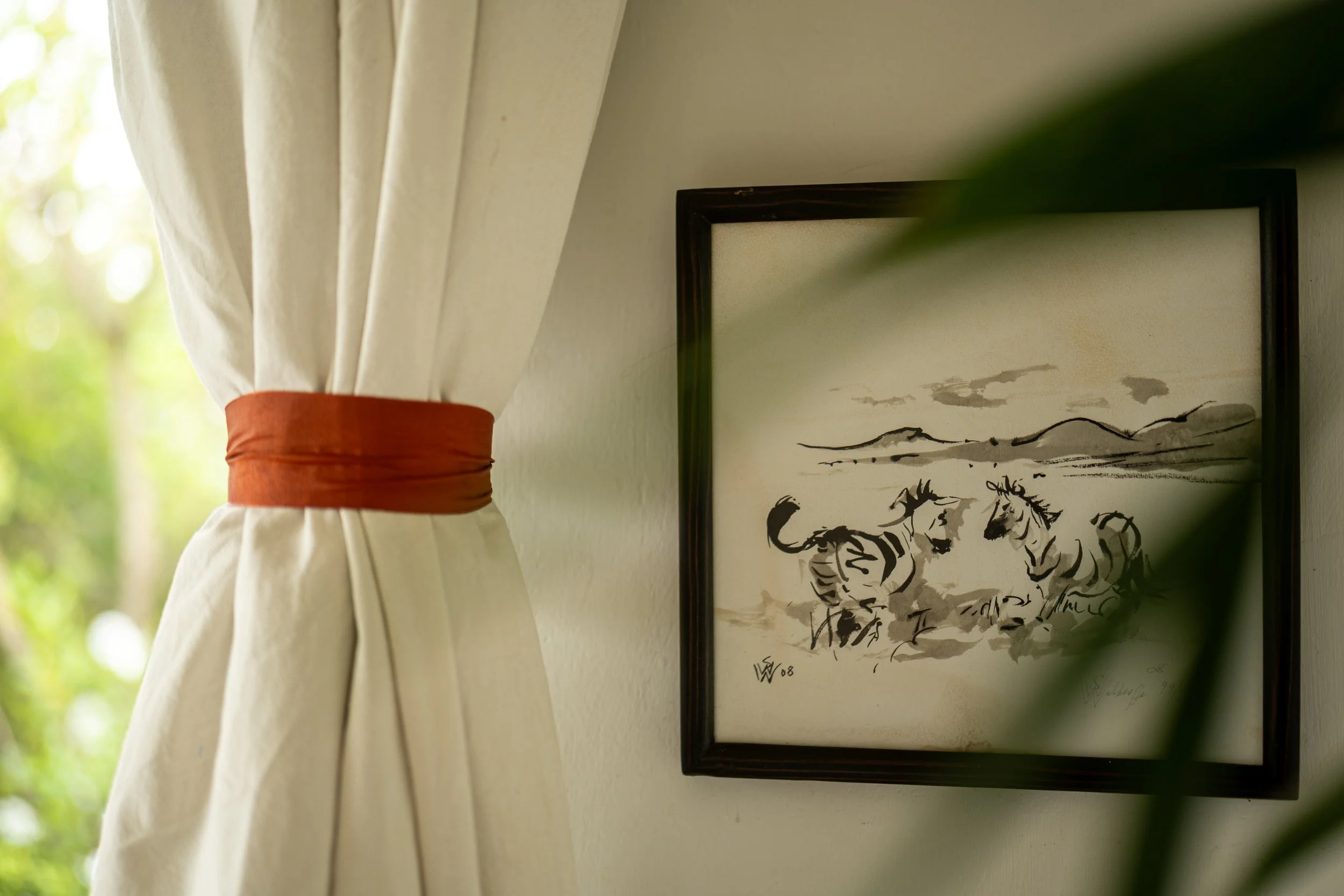 A framed ink drawing of horses and a landscape hanging on a wall, partially obscured by a white curtain with an orange tieback, with greenery visible outside a window.